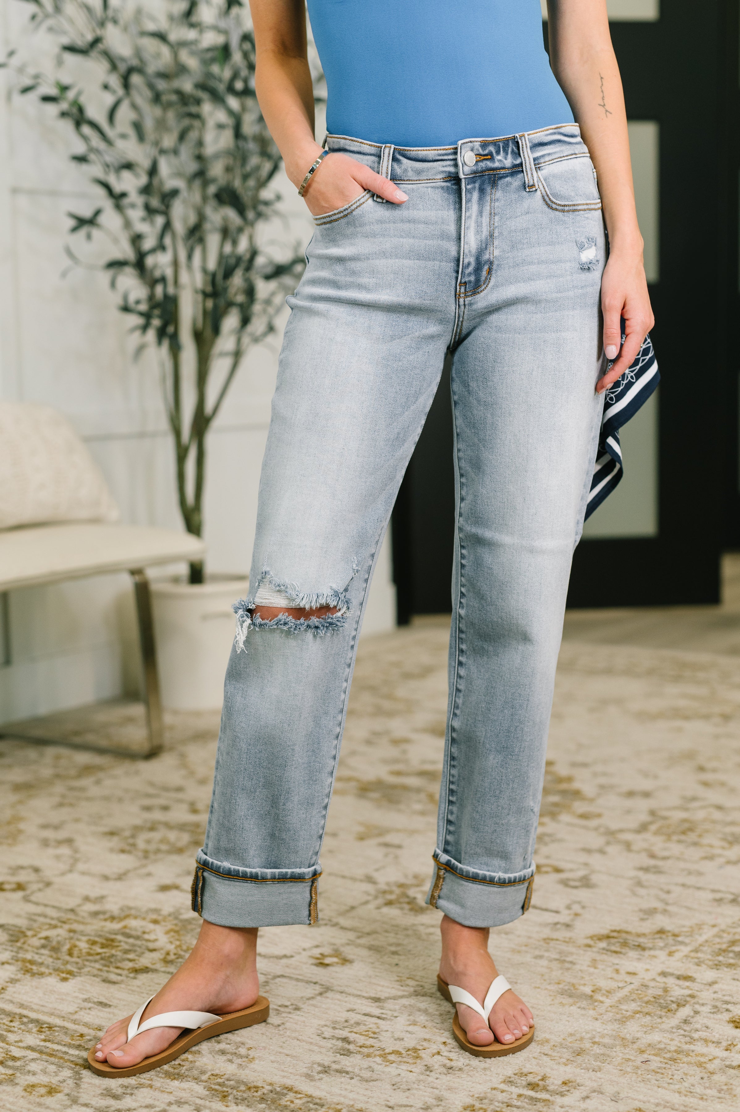 Woman wearing light blue mid-rise boyfriend jeans with a bandana loop, rolled cuffs, and white sandals indoors