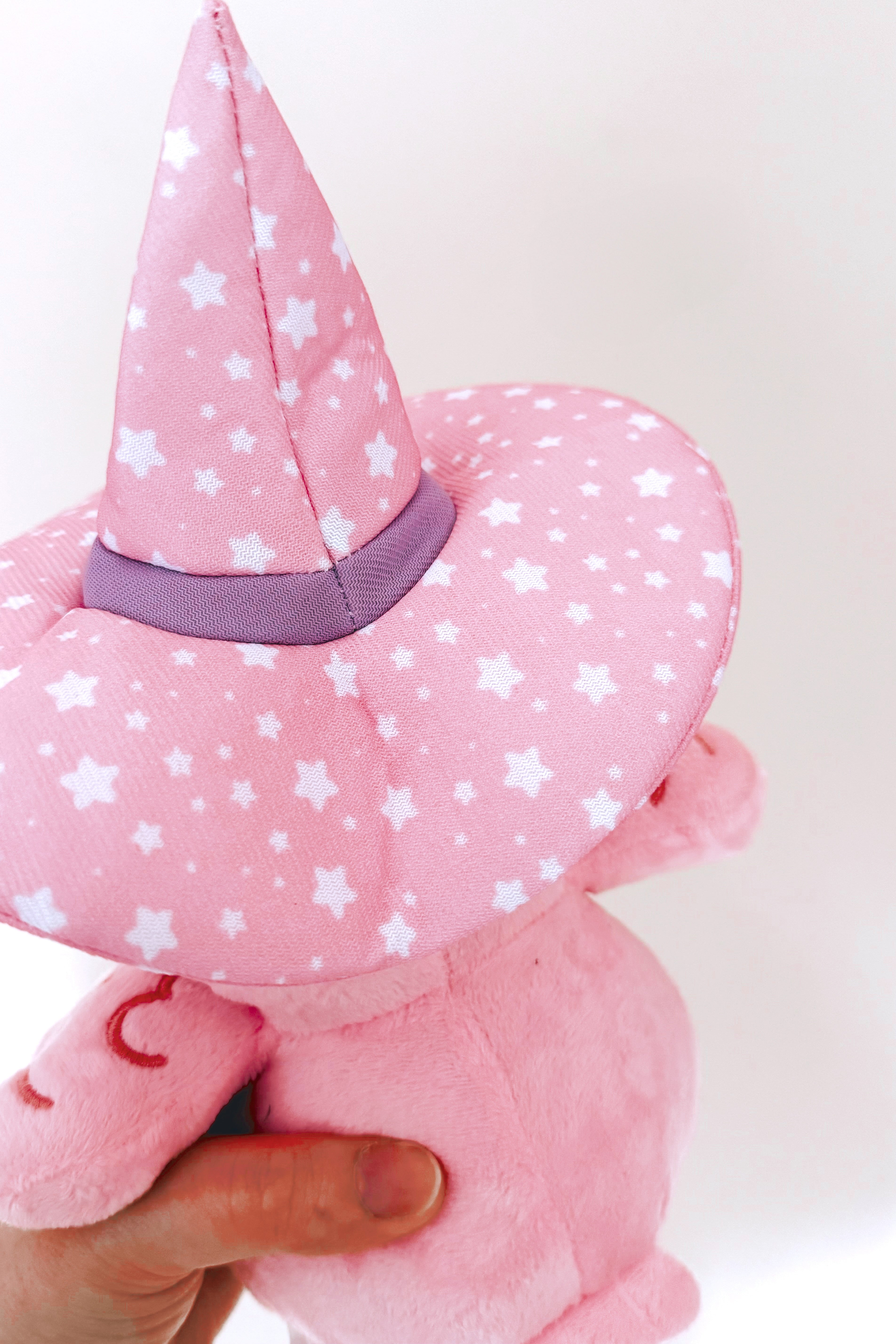 Close-up rear view of pink plushie wearing a star-patterned witch hat with purple band held by hand against white background