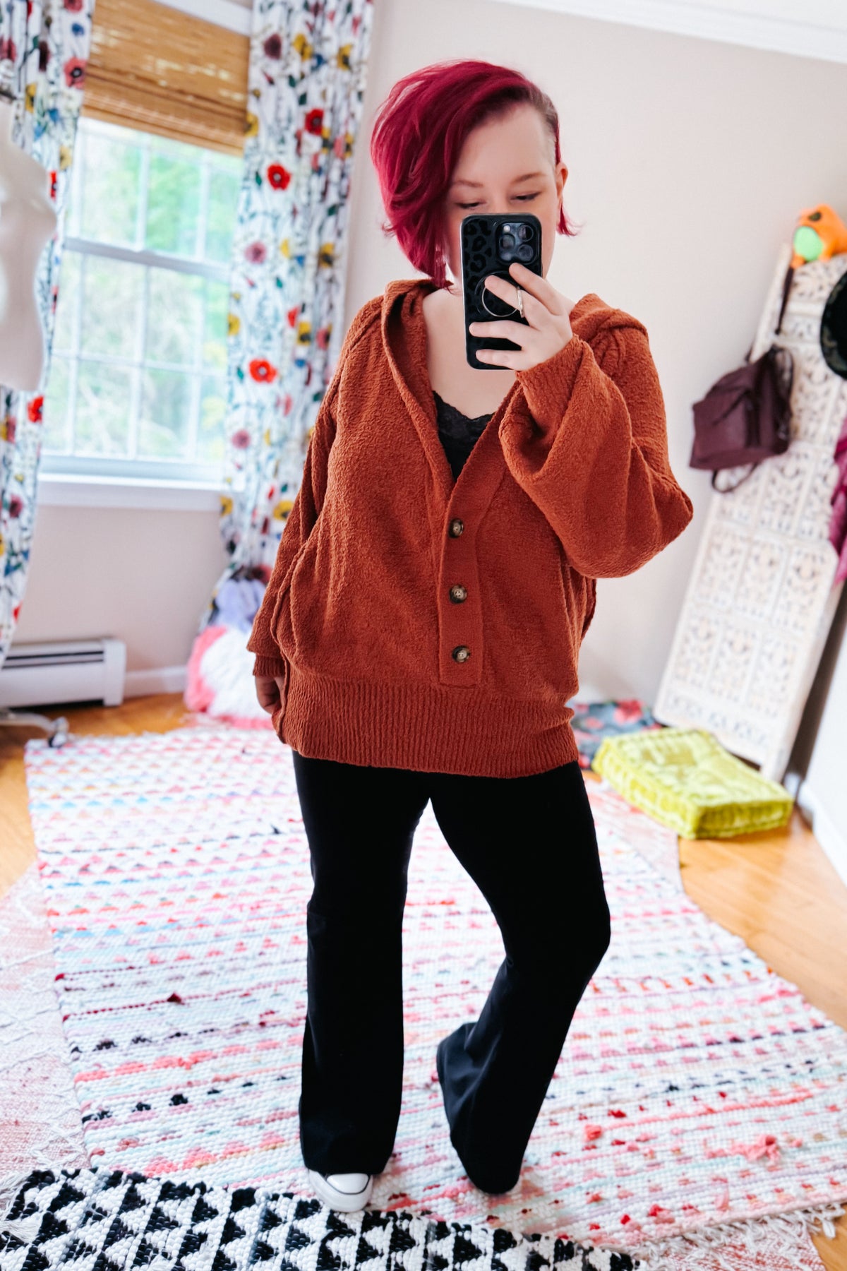 Woman taking a mirror selfie in a rust textured hooded henley sweater with half button closure black pants and white shoes indoors