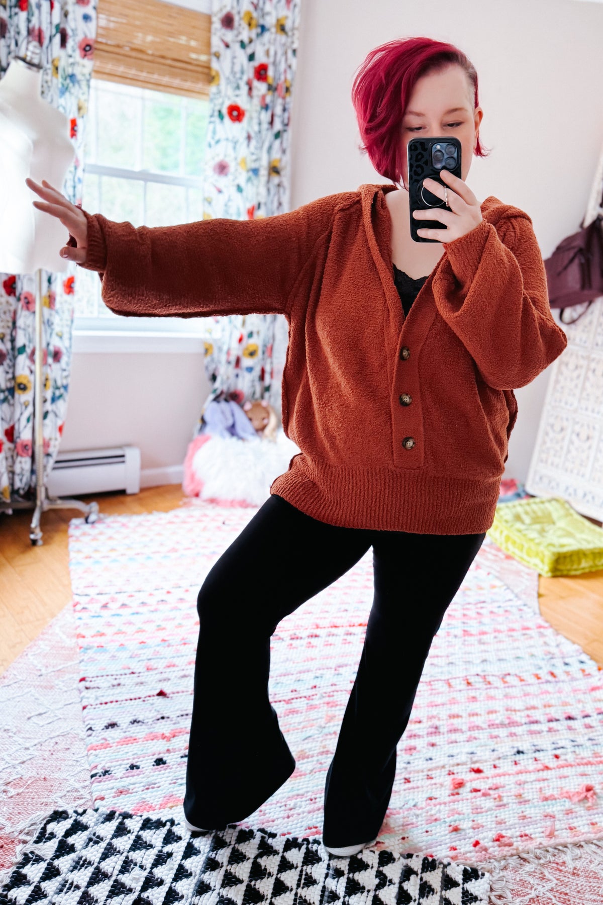 Woman taking a mirror selfie in a rust textured hooded henley sweater with half button closures and black pants indoors