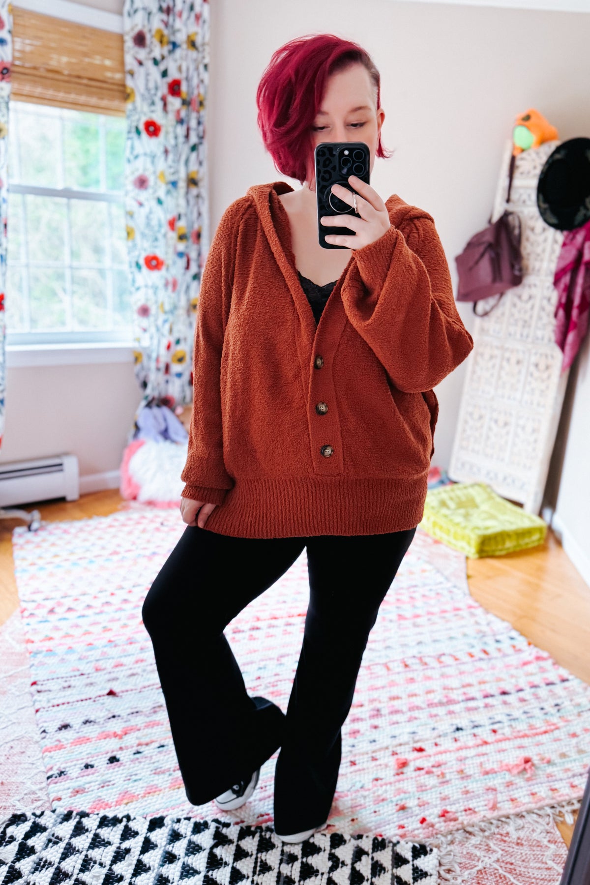 Woman taking a mirror selfie in a Rust textured knit hooded sweater with V neckline and button front styled with black pants indoors