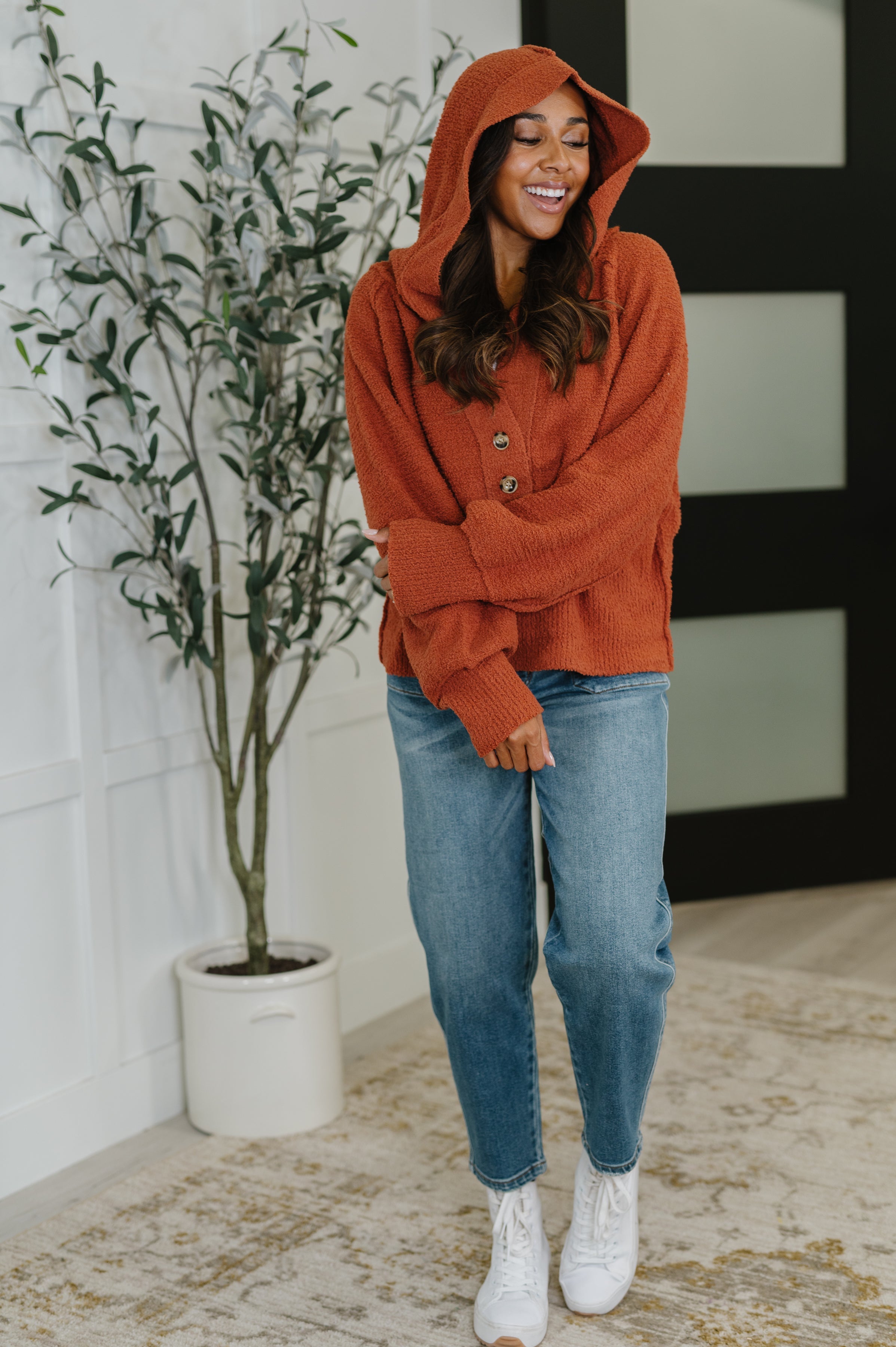 Smiling woman wearing rust-colored All For Me Henley Sweater with hood and buttons standing indoors by potted plant