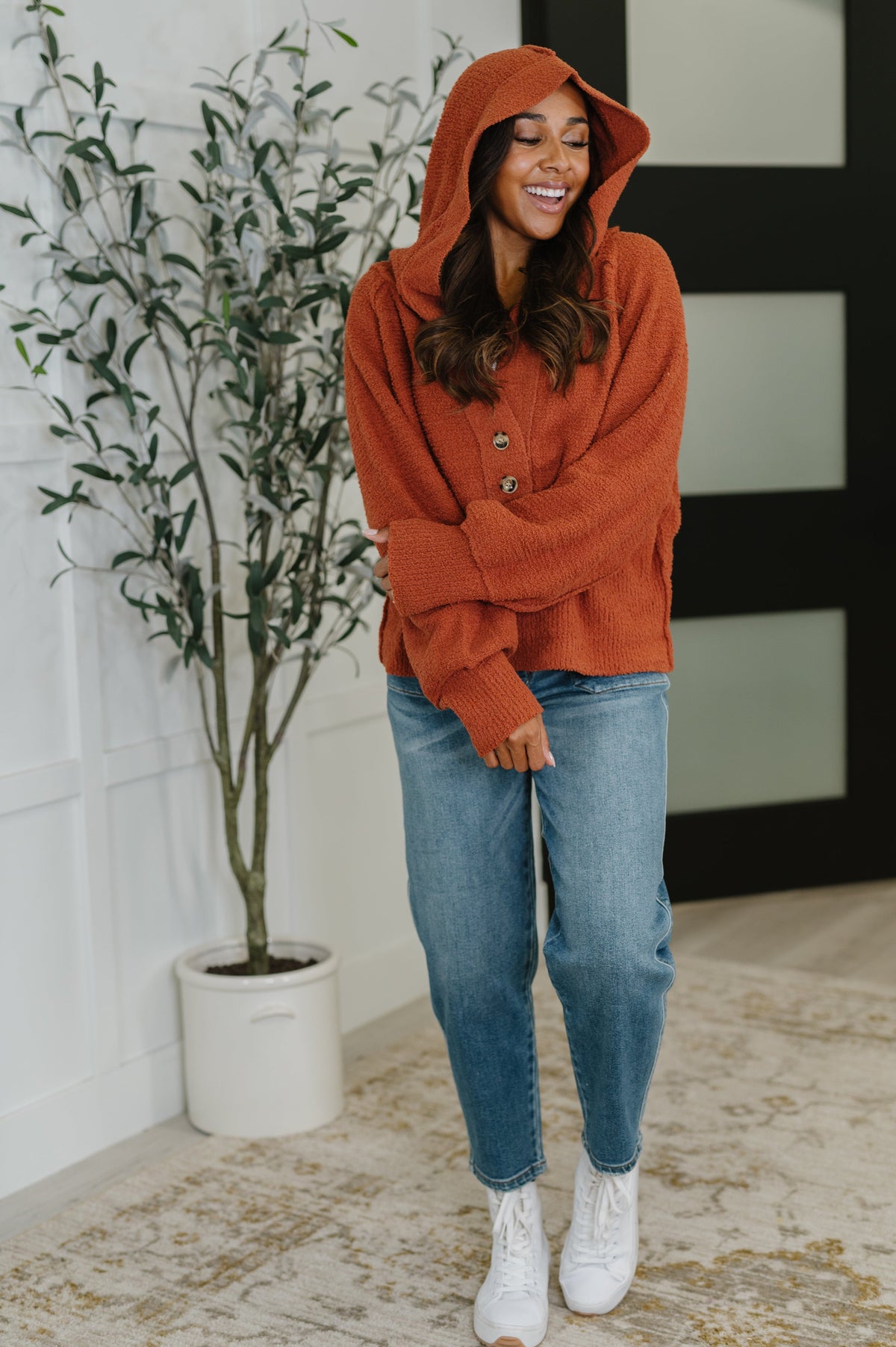 Woman smiling in a rust textured hooded henley sweater with half button closures blue jeans and white ankle boots indoors