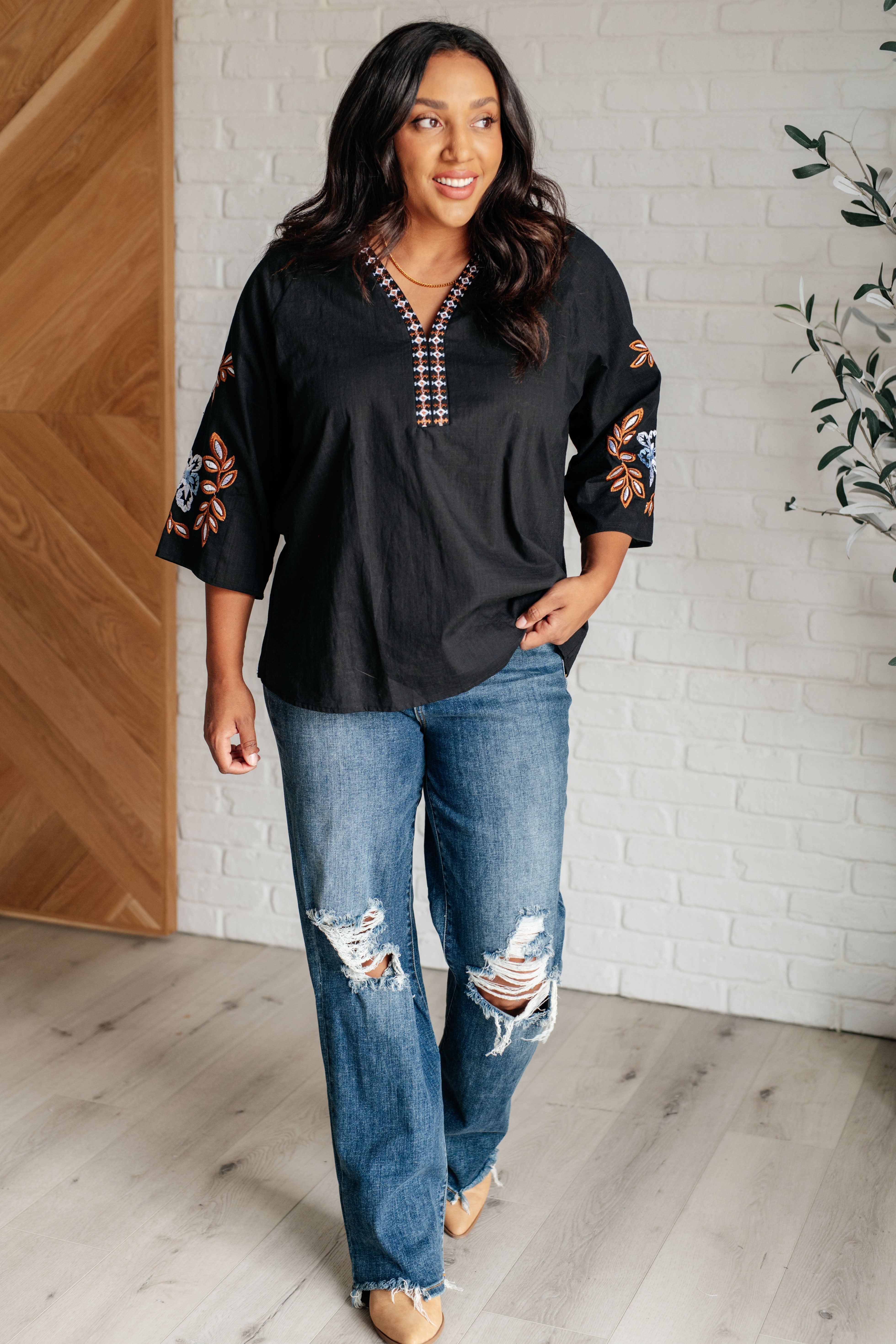Woman wearing a black embroidered v-neck blouse, blue jeans and tan boots standing against a white brick wall.