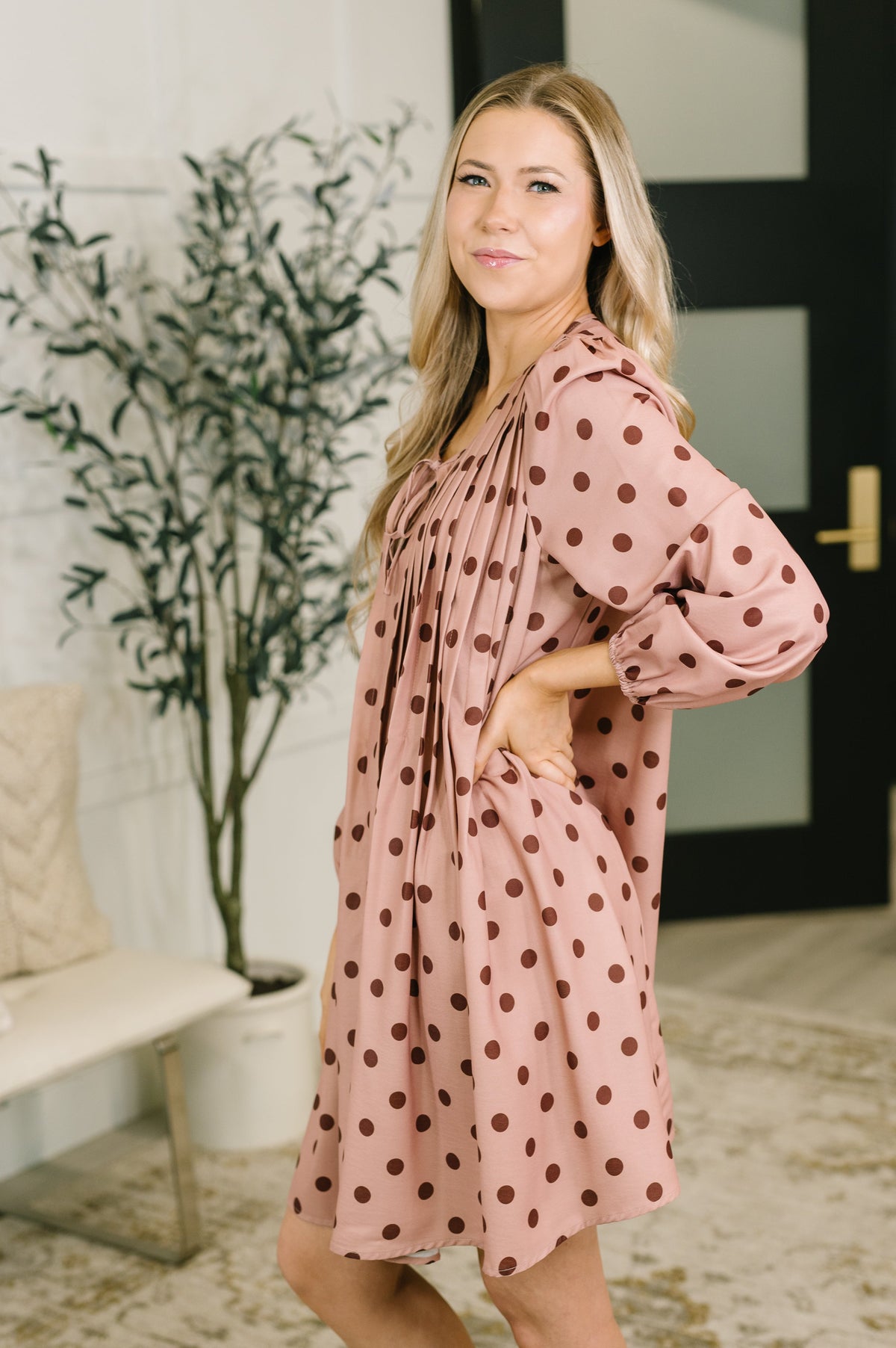 Woman models a pink pleated dress with brown polka dots and long sleeves in a bright modern interior