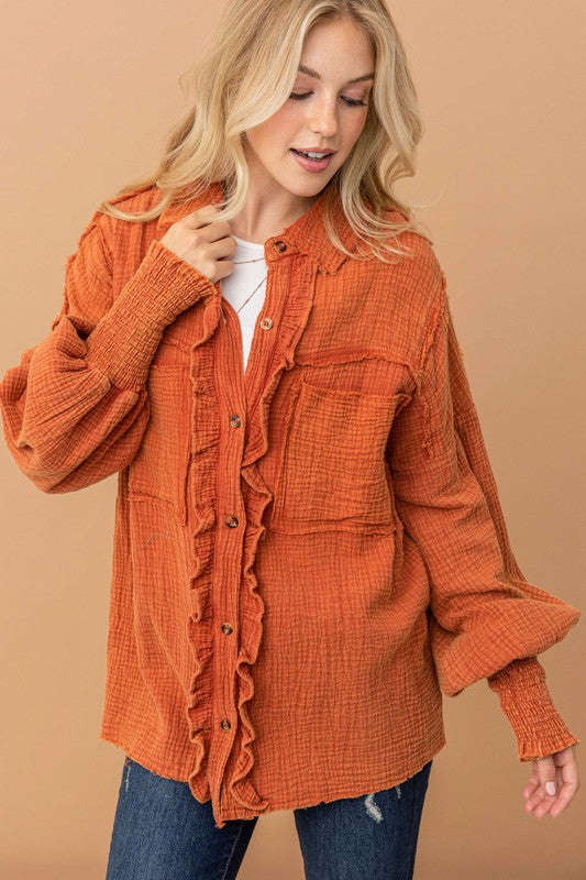 Blonde woman wearing textured burnt orange ruffled button-up shirt with smocked cuffs and chest pockets against tan background