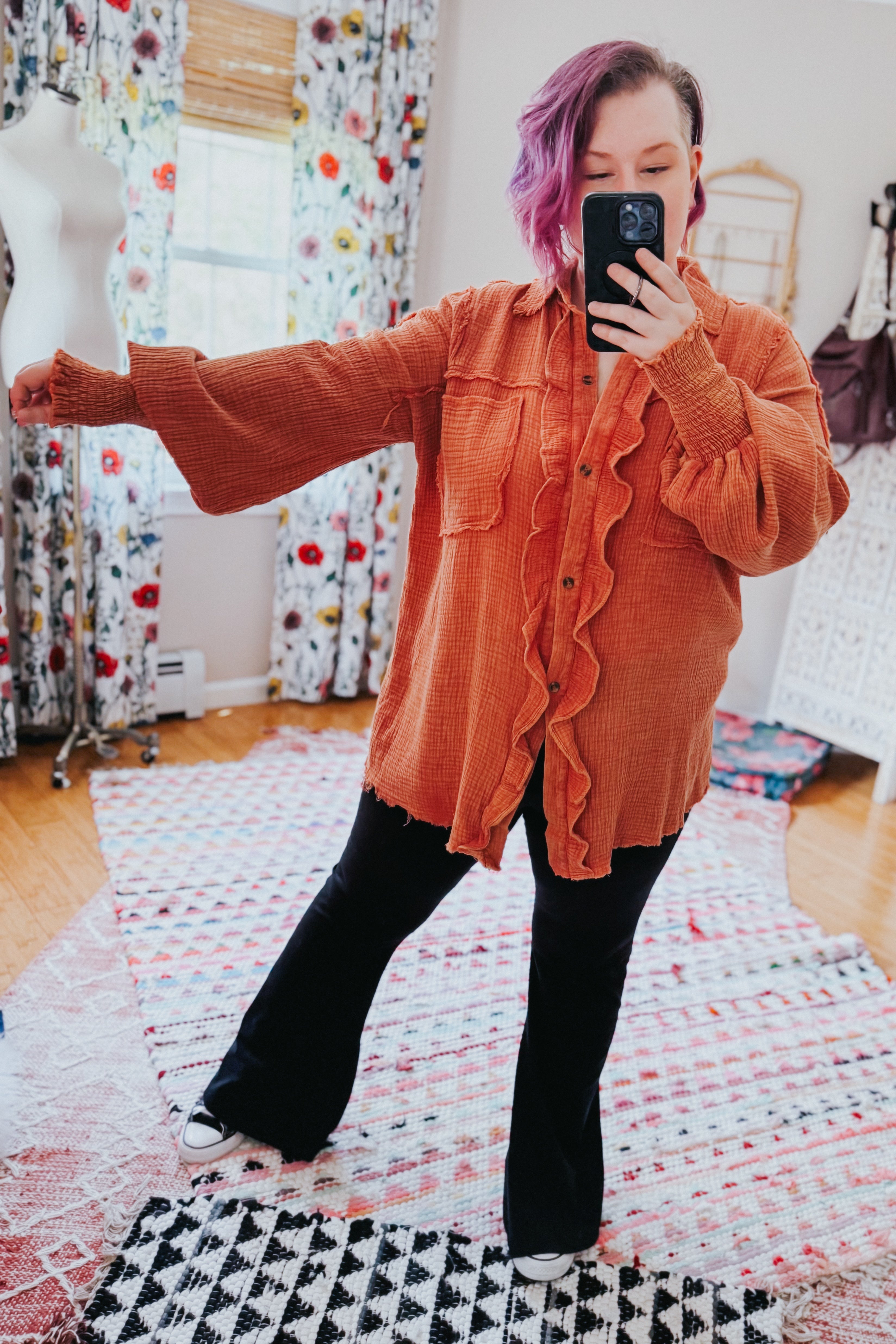 Woman with purple hair taking mirror selfie wearing loose orange ruffled button-up shirt with extended sleeves indoors