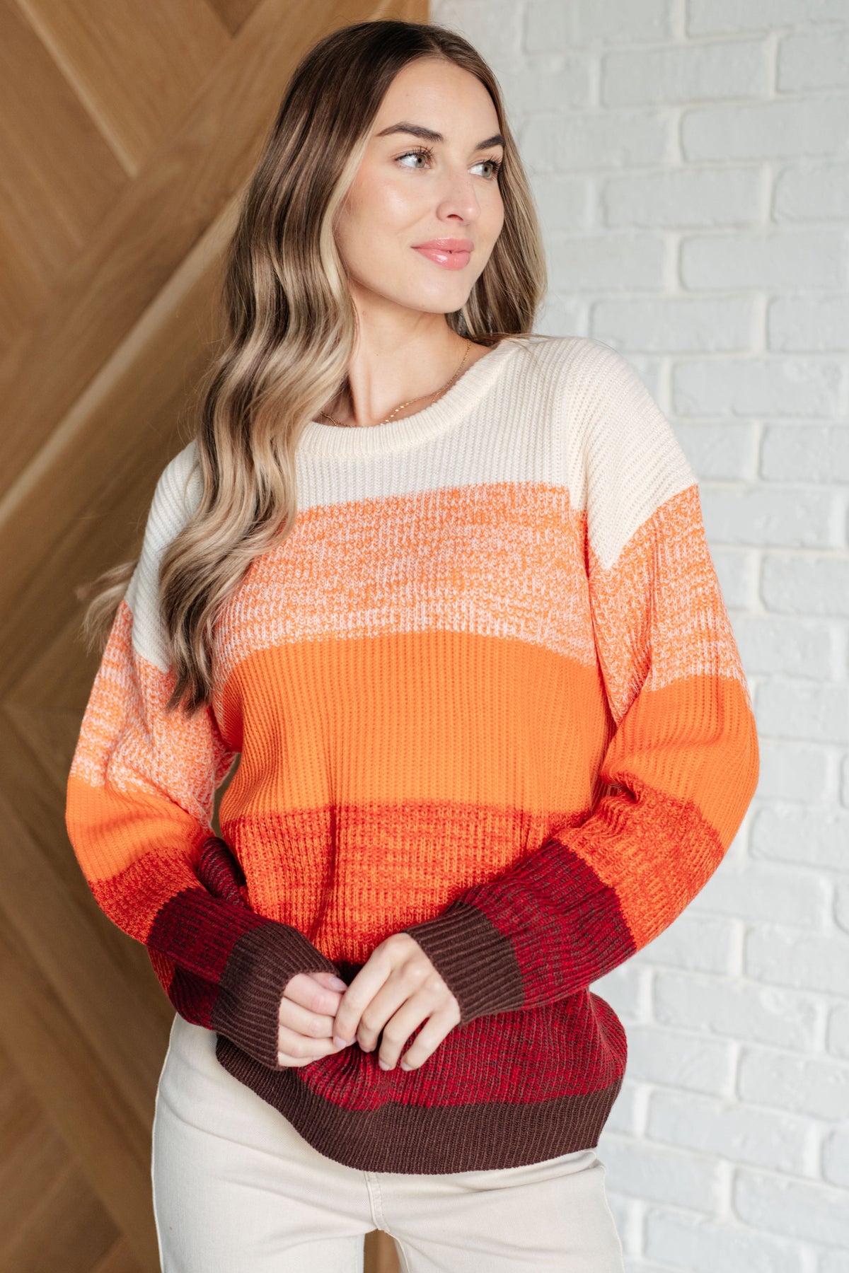 Woman wearing an orange color block sweater with cream and burgundy bands ribbed crew neckline and relaxed sleeves indoors