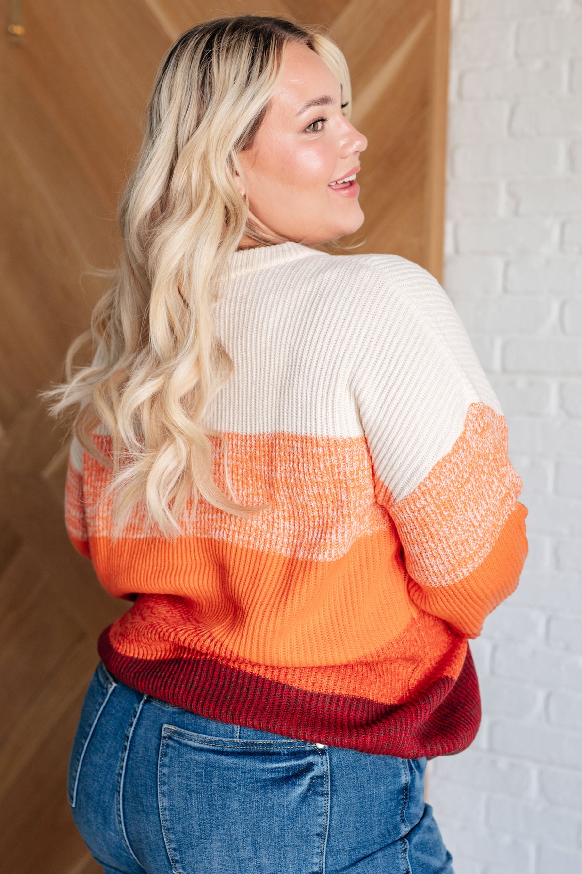 Woman showing the back of an orange color block sweater with cream and burgundy bands and relaxed sleeves styled with blue jeans indoors