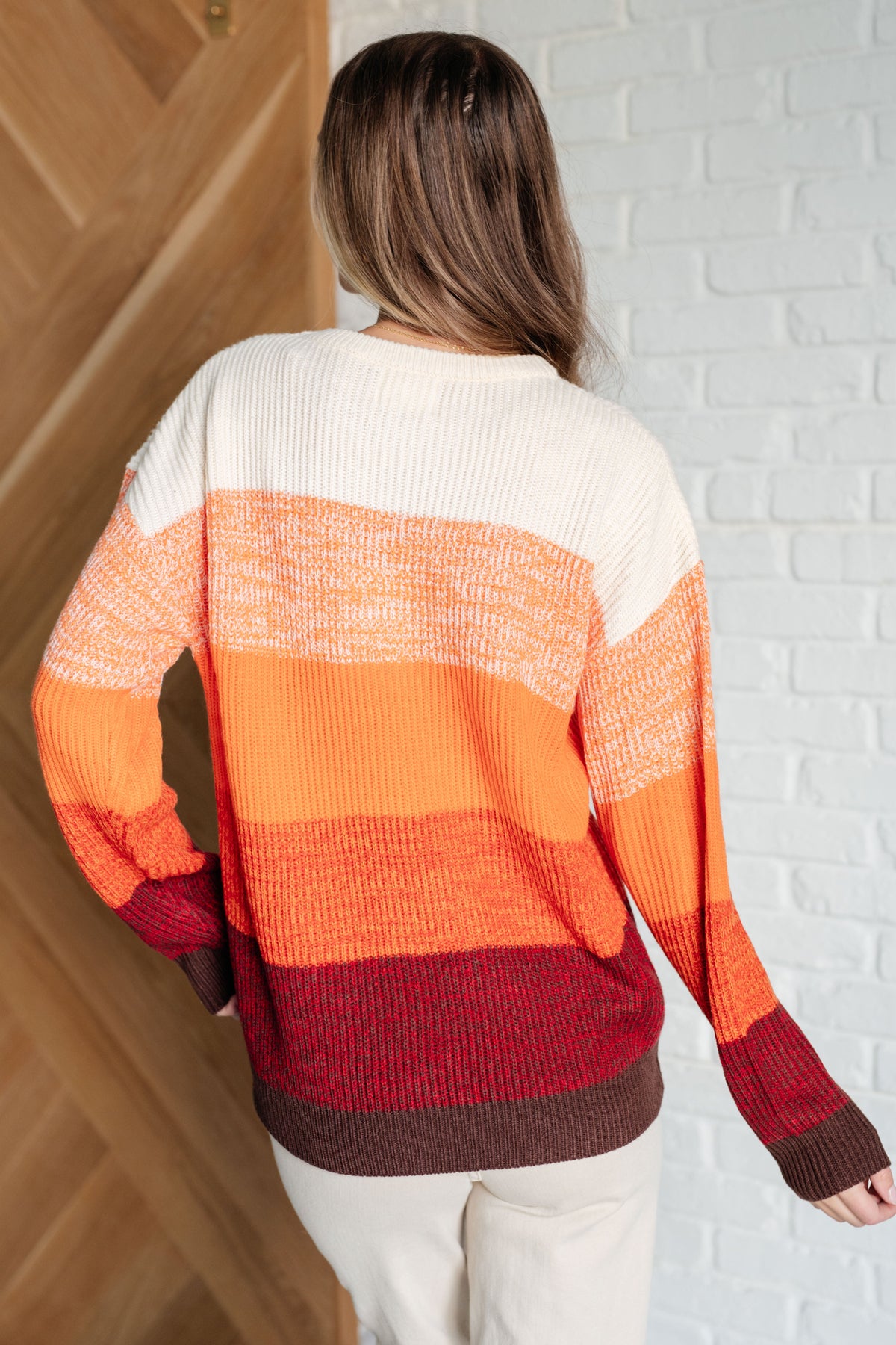 Back view of a woman wearing an orange color block sweater with cream and burgundy bands and relaxed drop shoulders styled with white pants indoors