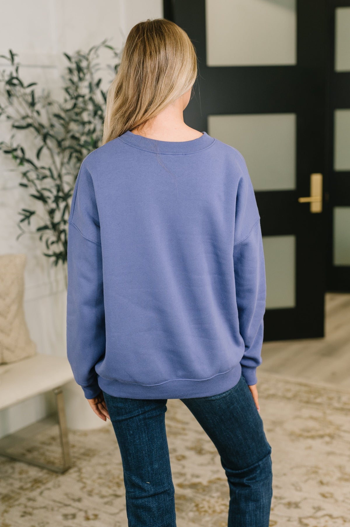 Woman seen from behind wearing a relaxed blue pullover with drop shoulders and jeans in a softly styled home interior