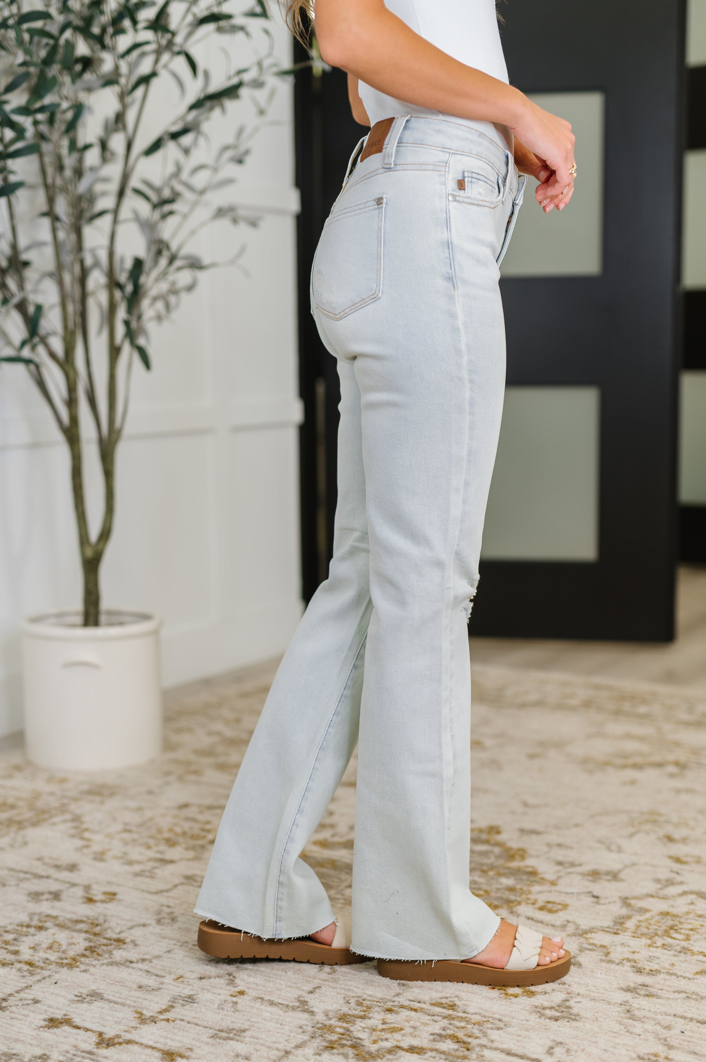 Side profile of light wash Alyse Mid Rise Destroy & Fray Hem Bootcut jeans with frayed edges and sandals indoors