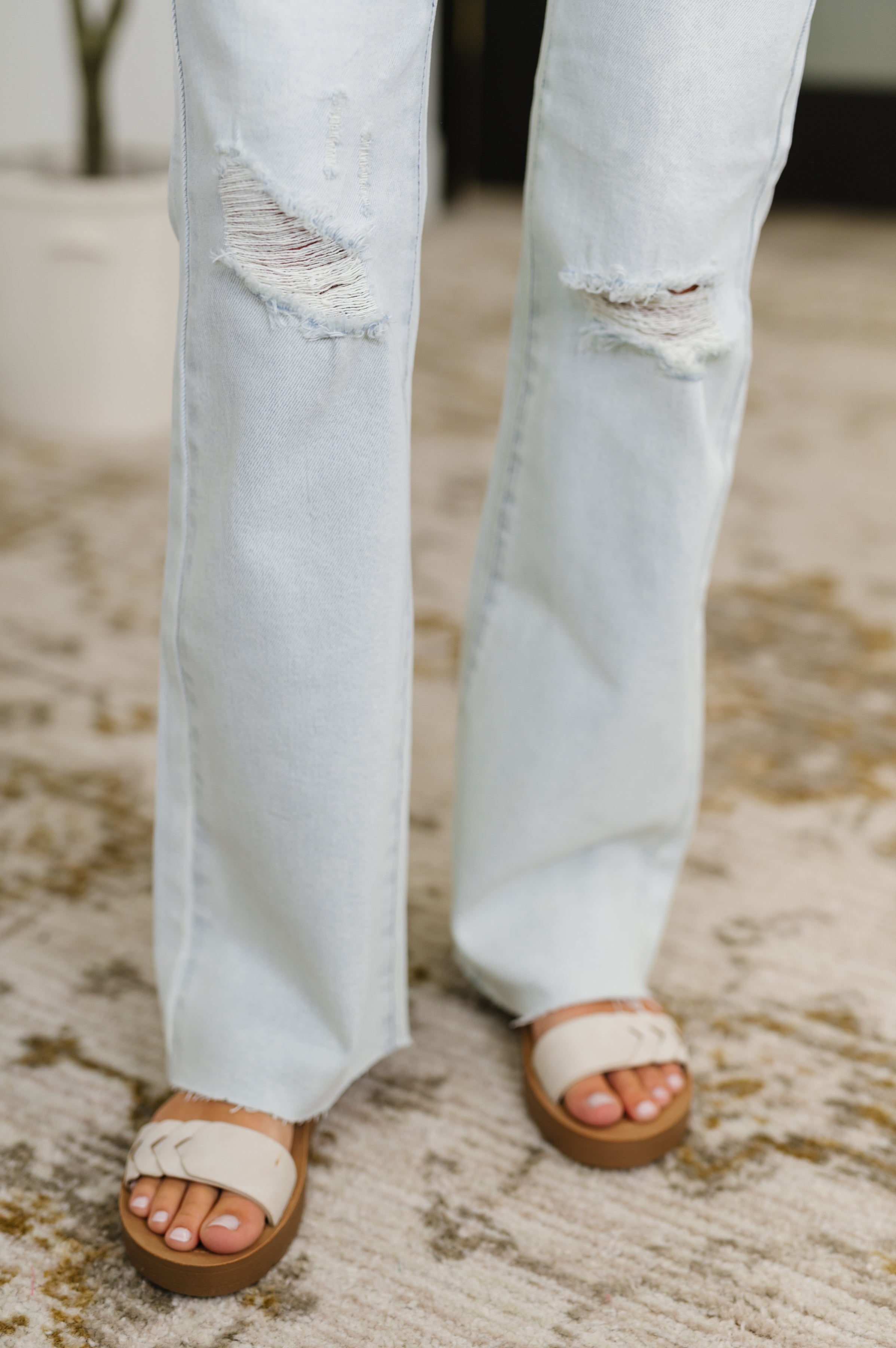 Close-up of light wash Alyse Mid Rise Destroy & Fray Hem Bootcut jeans with distressed knee details and sandals on carpet