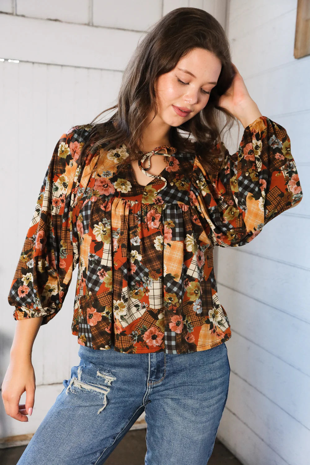 Woman wearing a floral patchwork print blouse and jeans against a white wall.