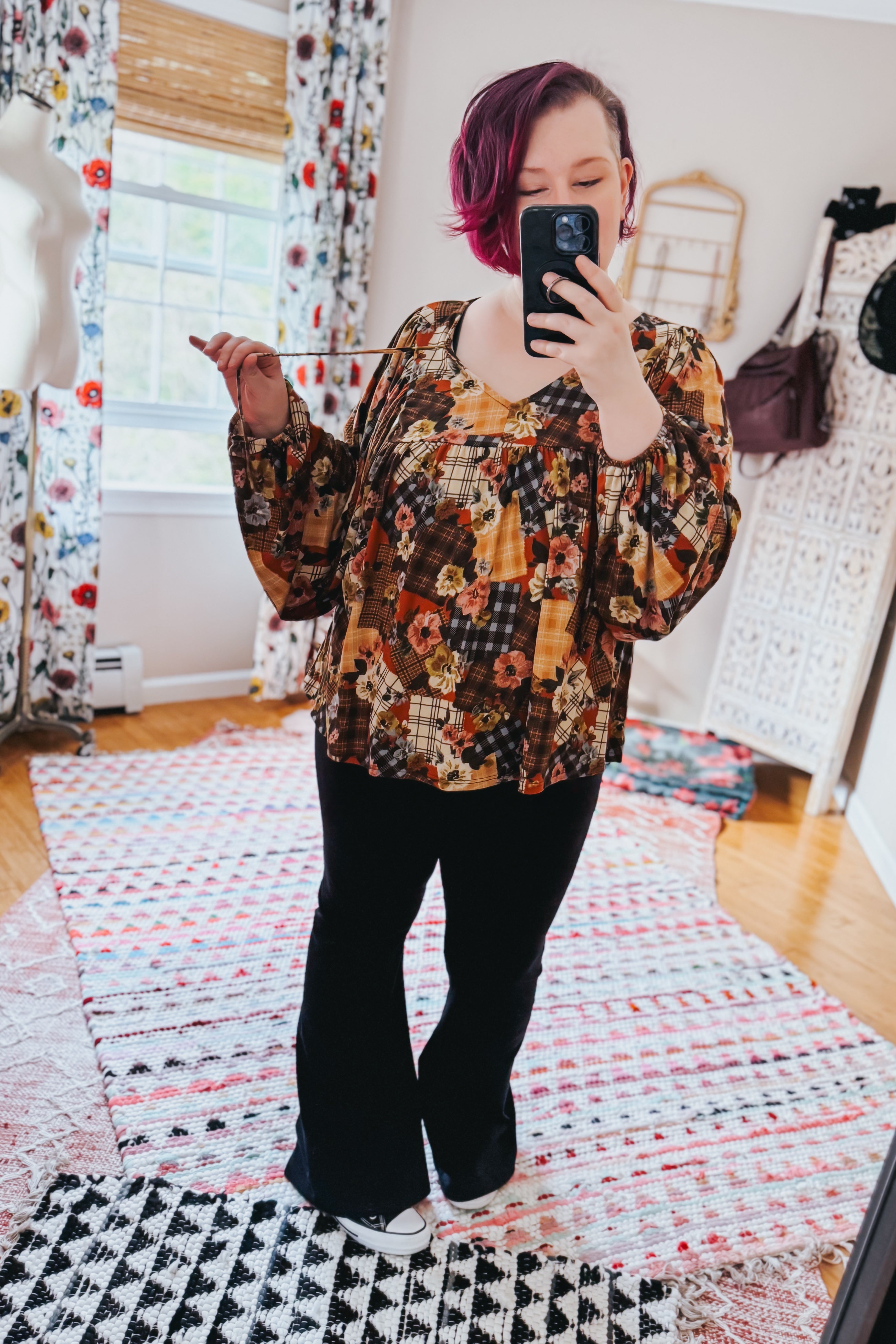 Woman taking a mirror selfie wearing a floral patchwork print blouse and black pants in a room with colorful curtains and a patterned rug.