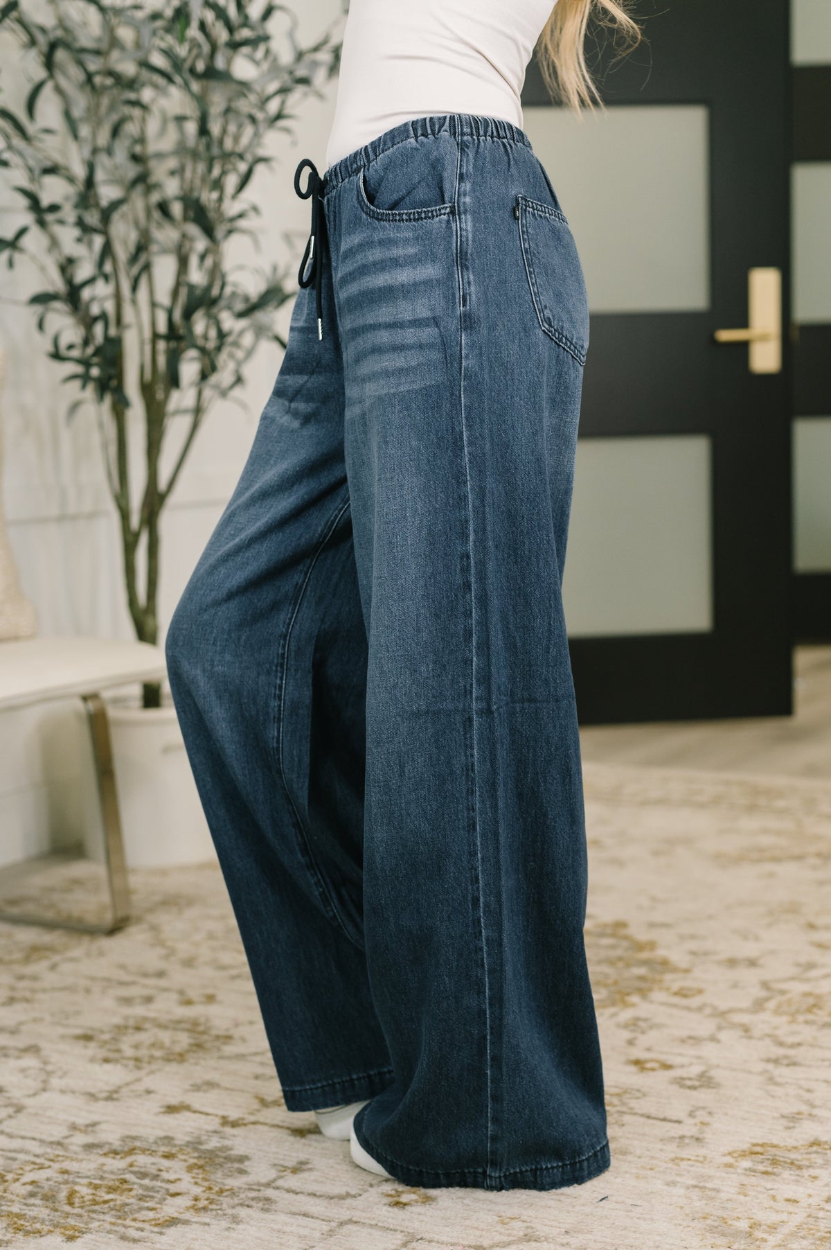 Side view of a woman wearing blue high waist palazzo jeans with an ultra wide leg silhouette in a modern home interior