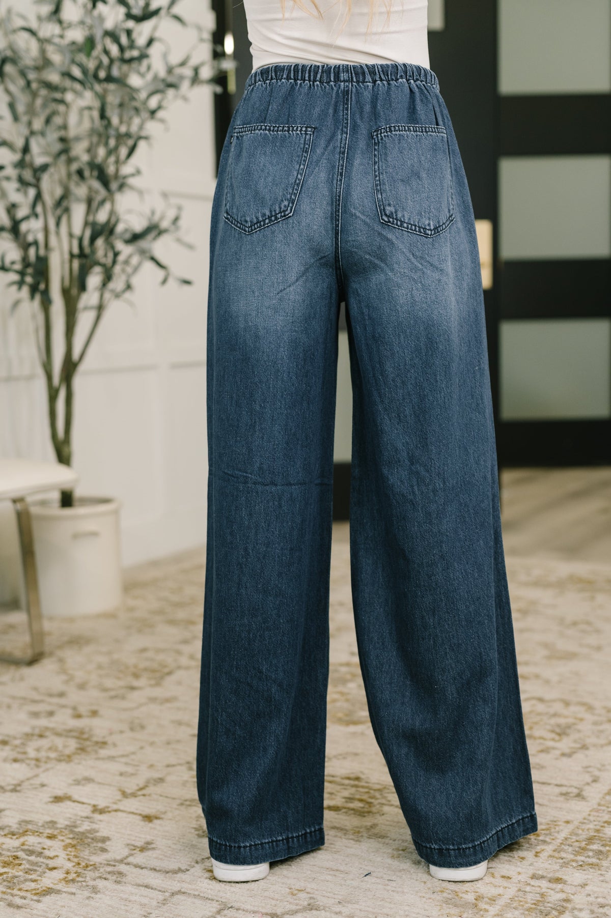 Back view of a woman wearing blue high-waisted wide-leg denim jeans with patch pockets and a white top indoors