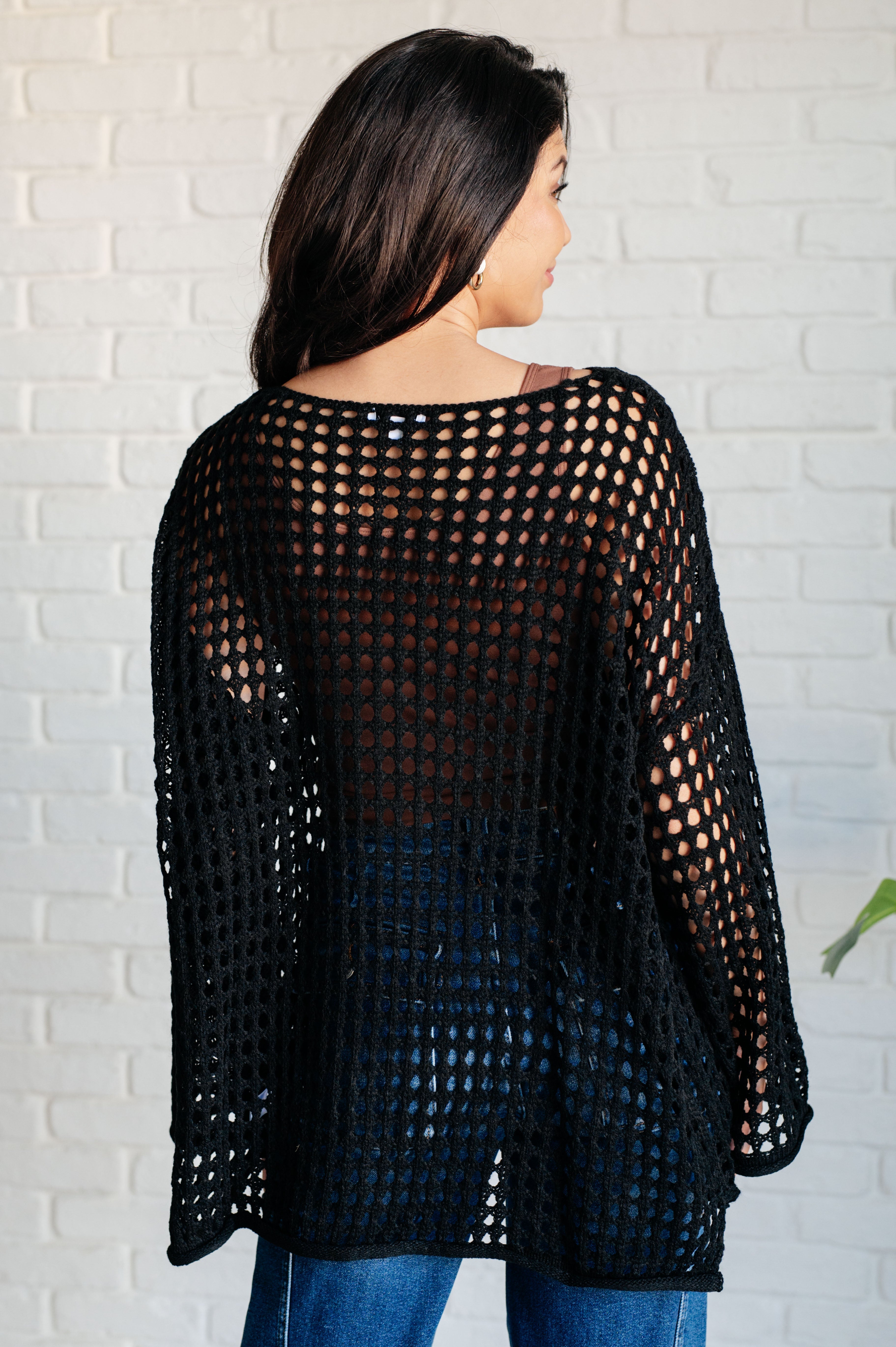 Woman wearing Ask Anyway black open mesh fishnet sweater viewed from back against white brick wall