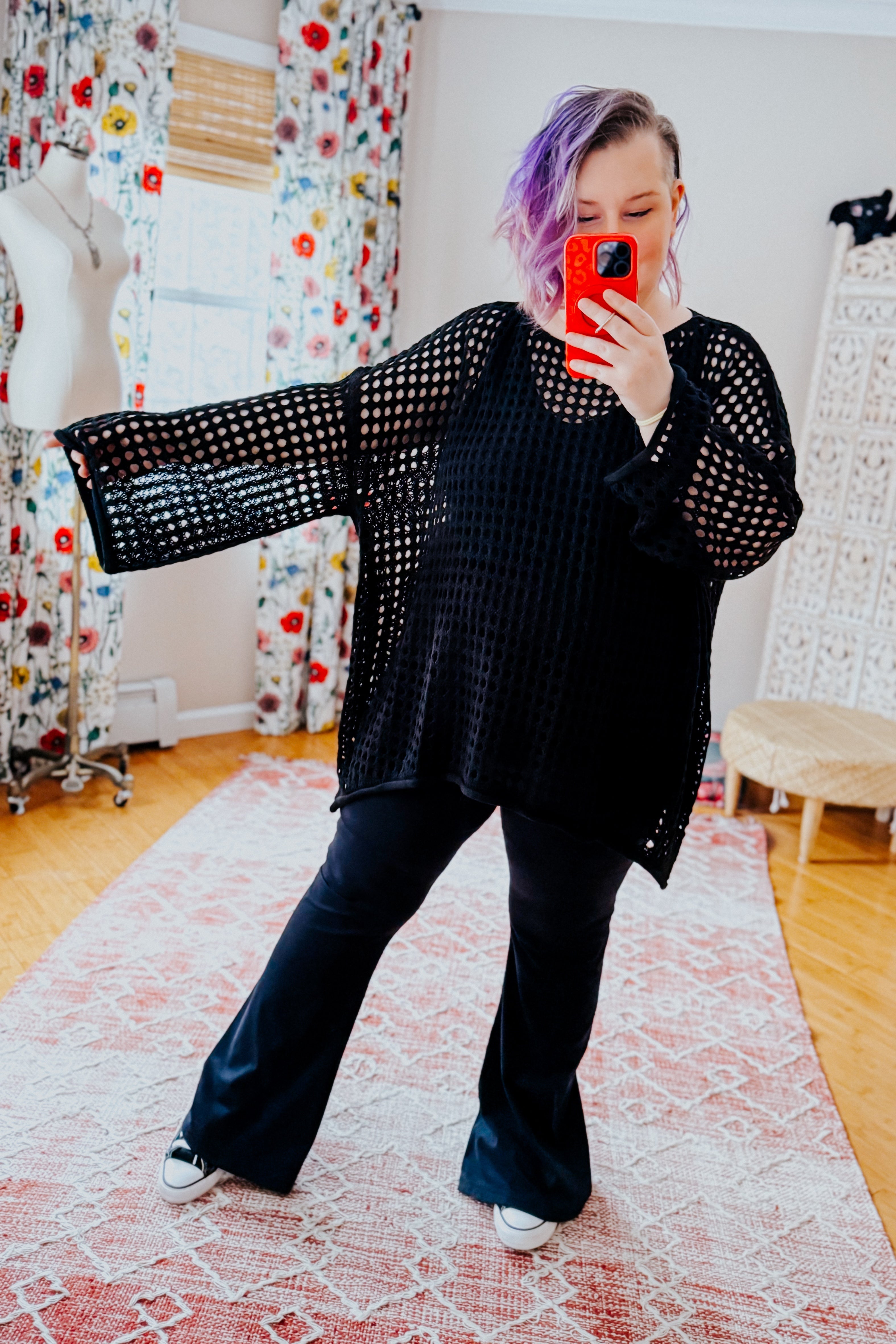 Person with purple hair modeling Ask Anyway oversized black fishnet sweater with wide sleeves and flared pants indoors