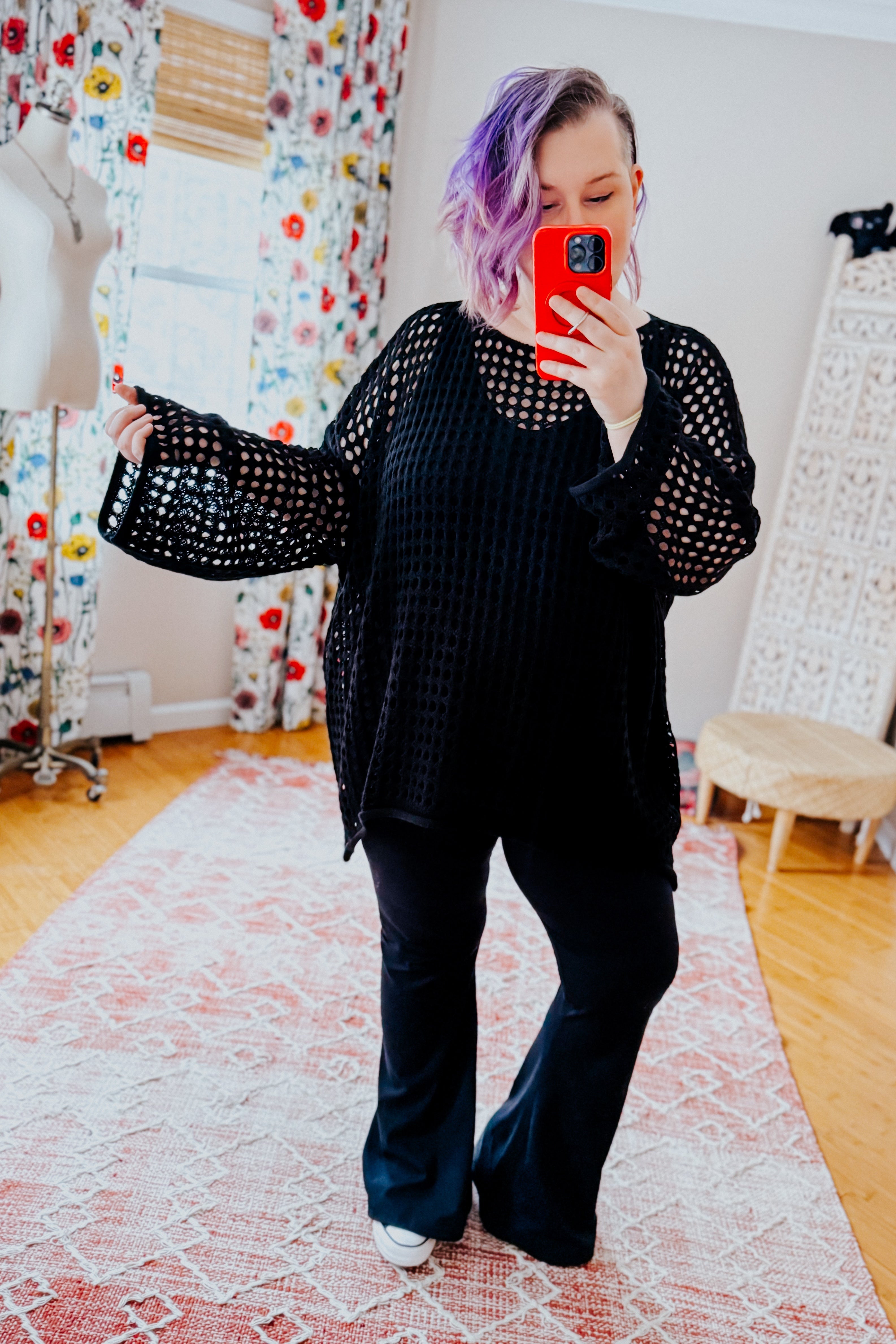 Woman with purple hair wearing Ask Anyway black fishnet sweater with wide sleeves taking red phone selfie indoors on floral rug
