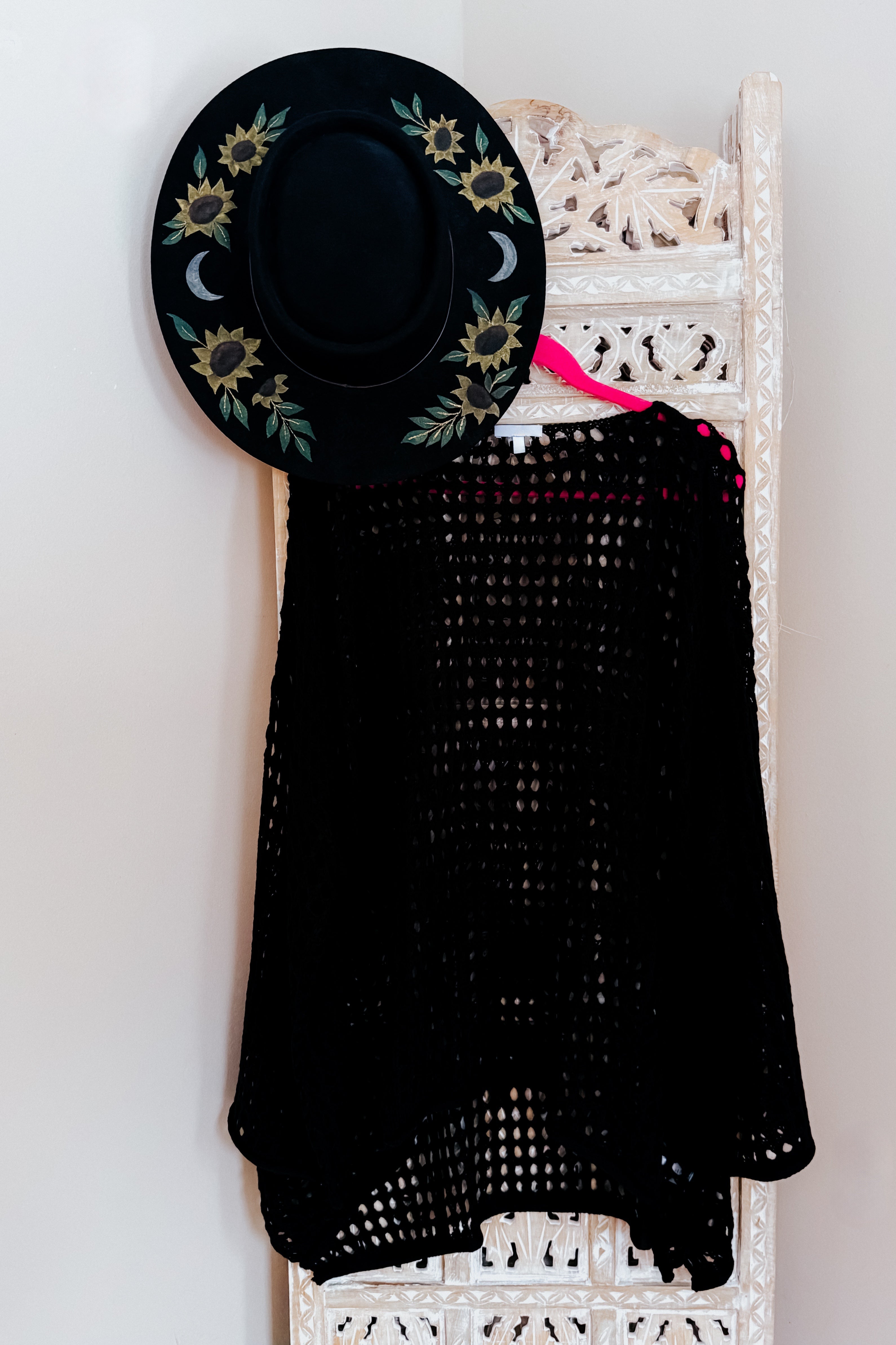 Black Ask Anyway fishnet sweater hanging on pink hanger on carved white wooden panel with sunflower moon decorated black hat