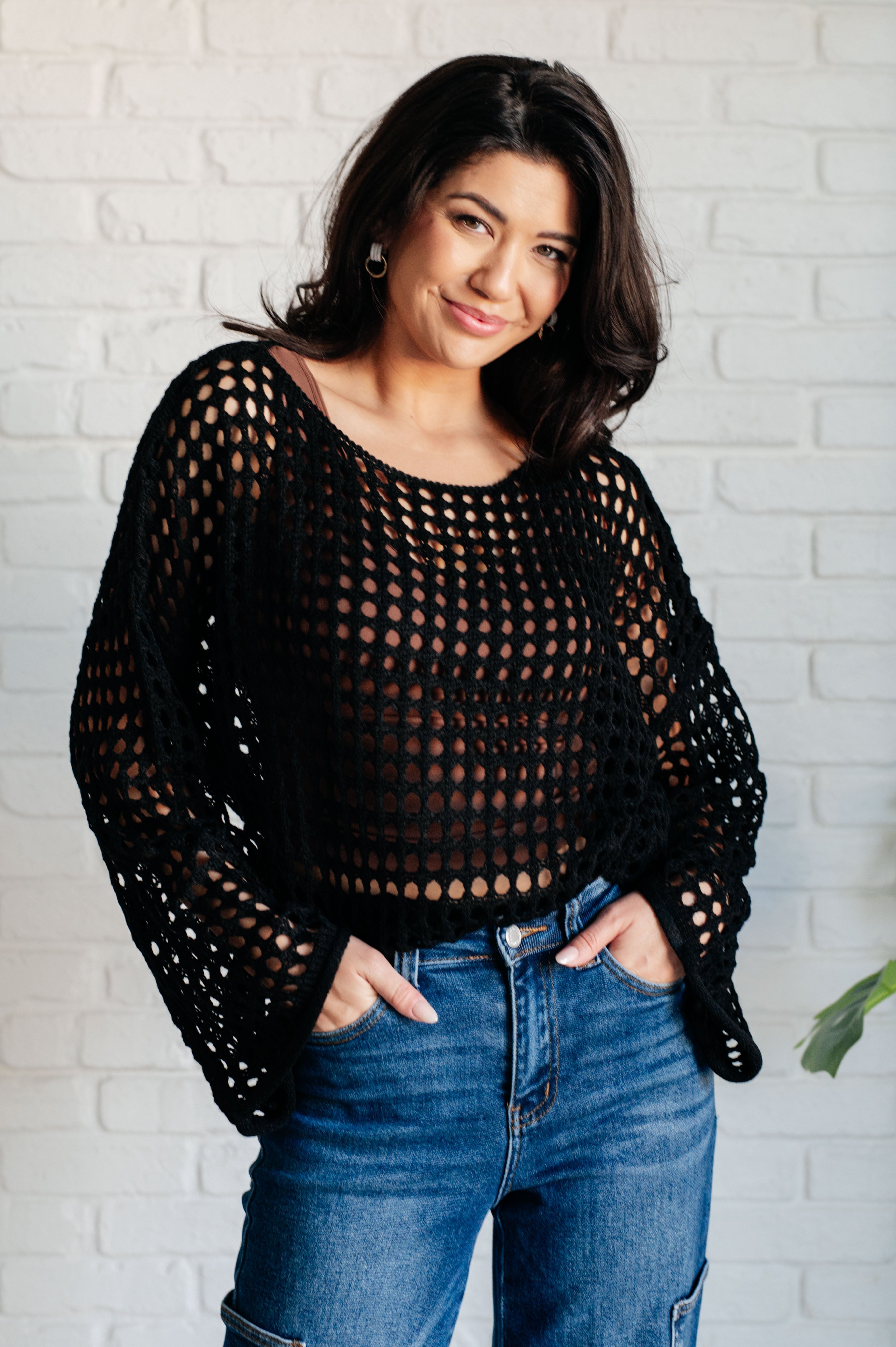 Smiling woman wearing Ask Anyway black fishnet sweater with wide sleeves paired with high-waisted blue jeans indoors