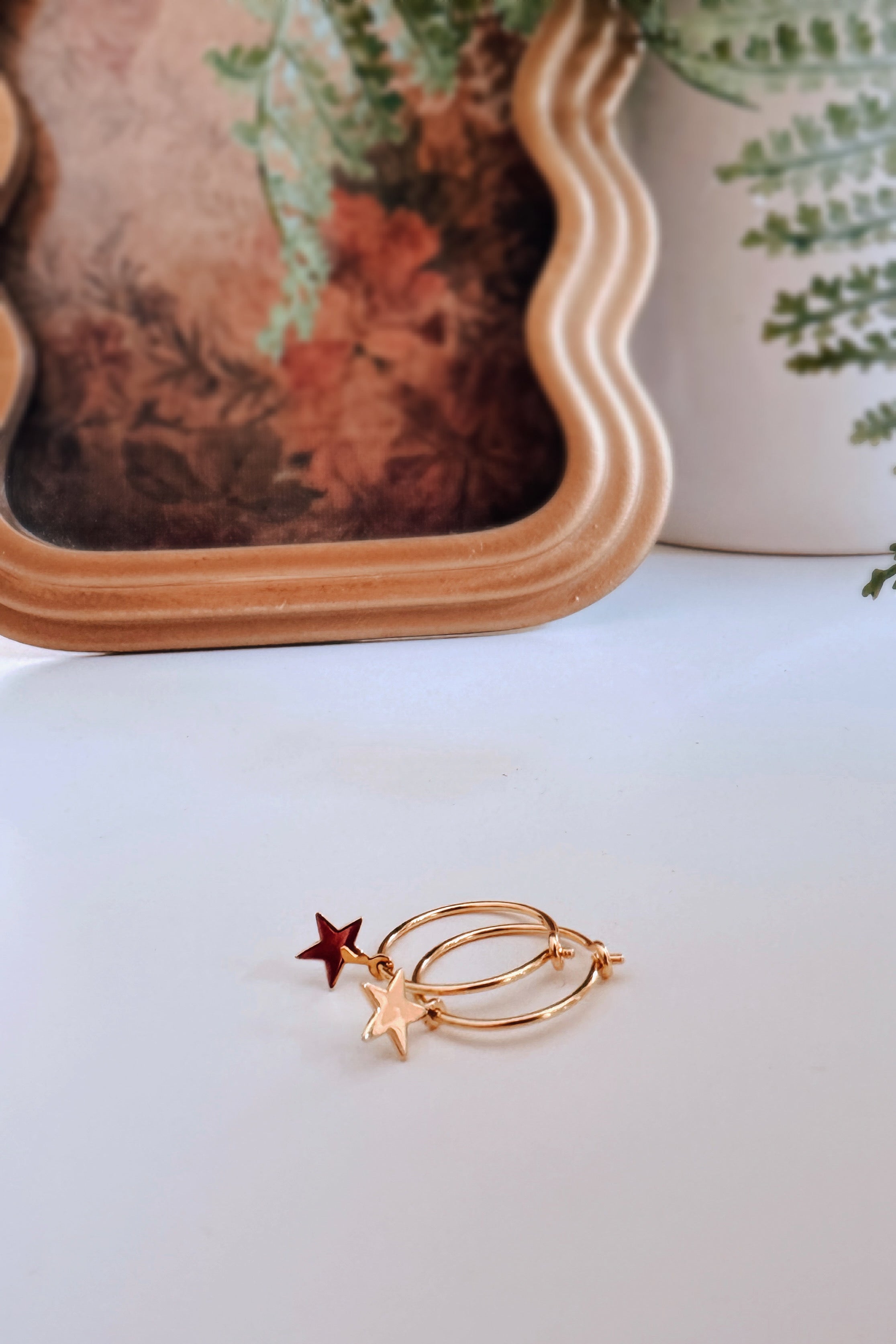 Pair of gold hoop earrings with red and gold star charms placed on white surface near wavy mirror and green plant leaves