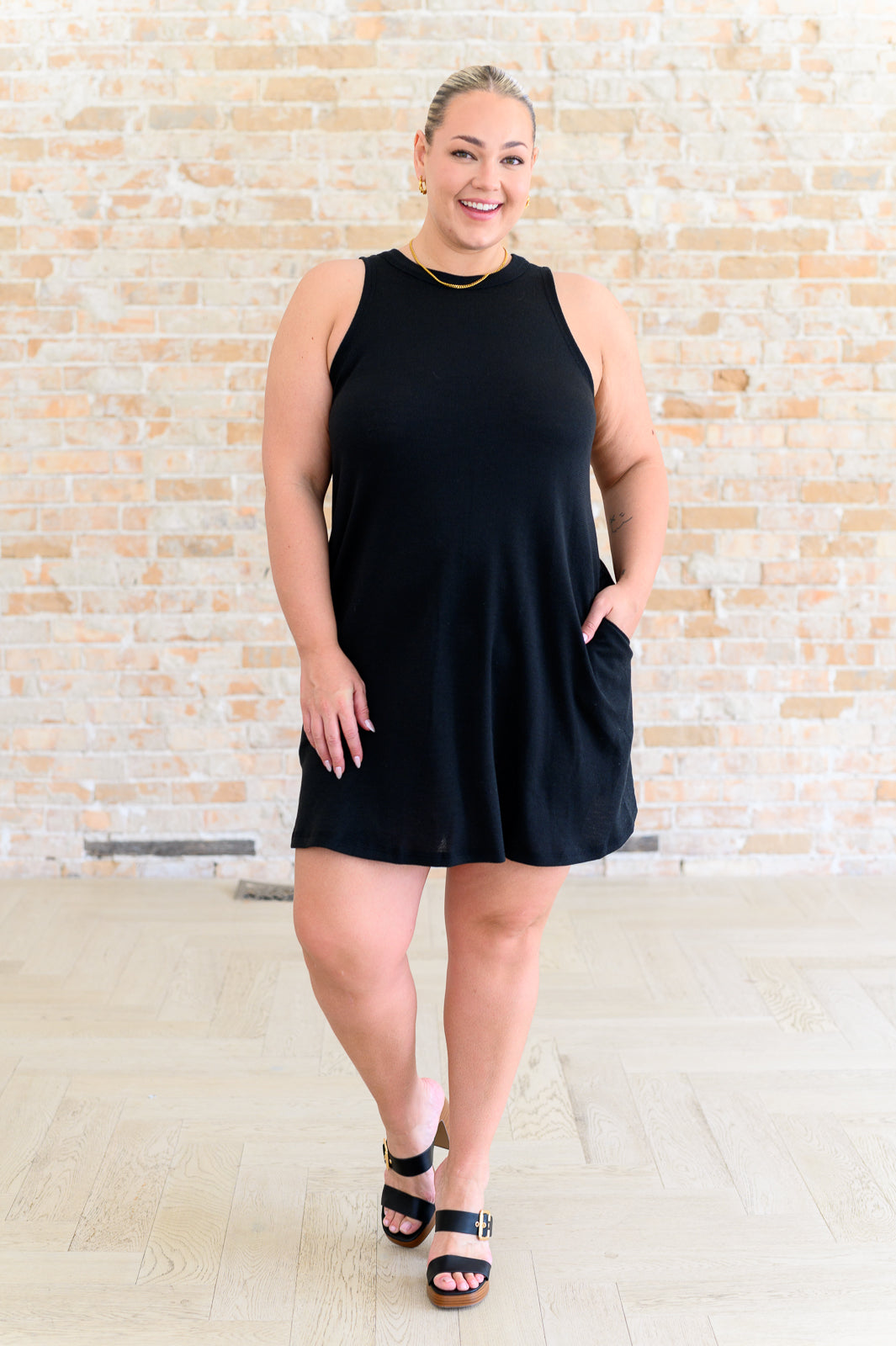 Smiling woman wearing a sleeveless black dress with pockets and gold jewelry standing indoors on light hardwood floor