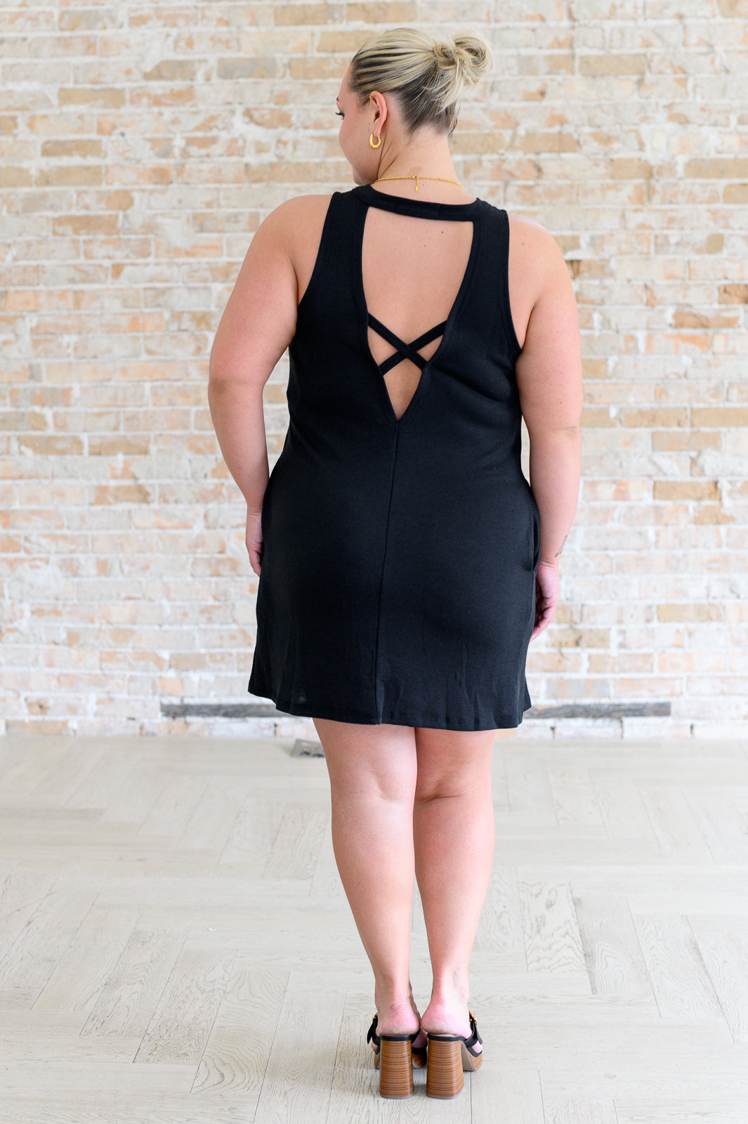 Plus-size woman wearing a black knit dress with crisscross cutout back and wooden block heel sandals against a brick wall