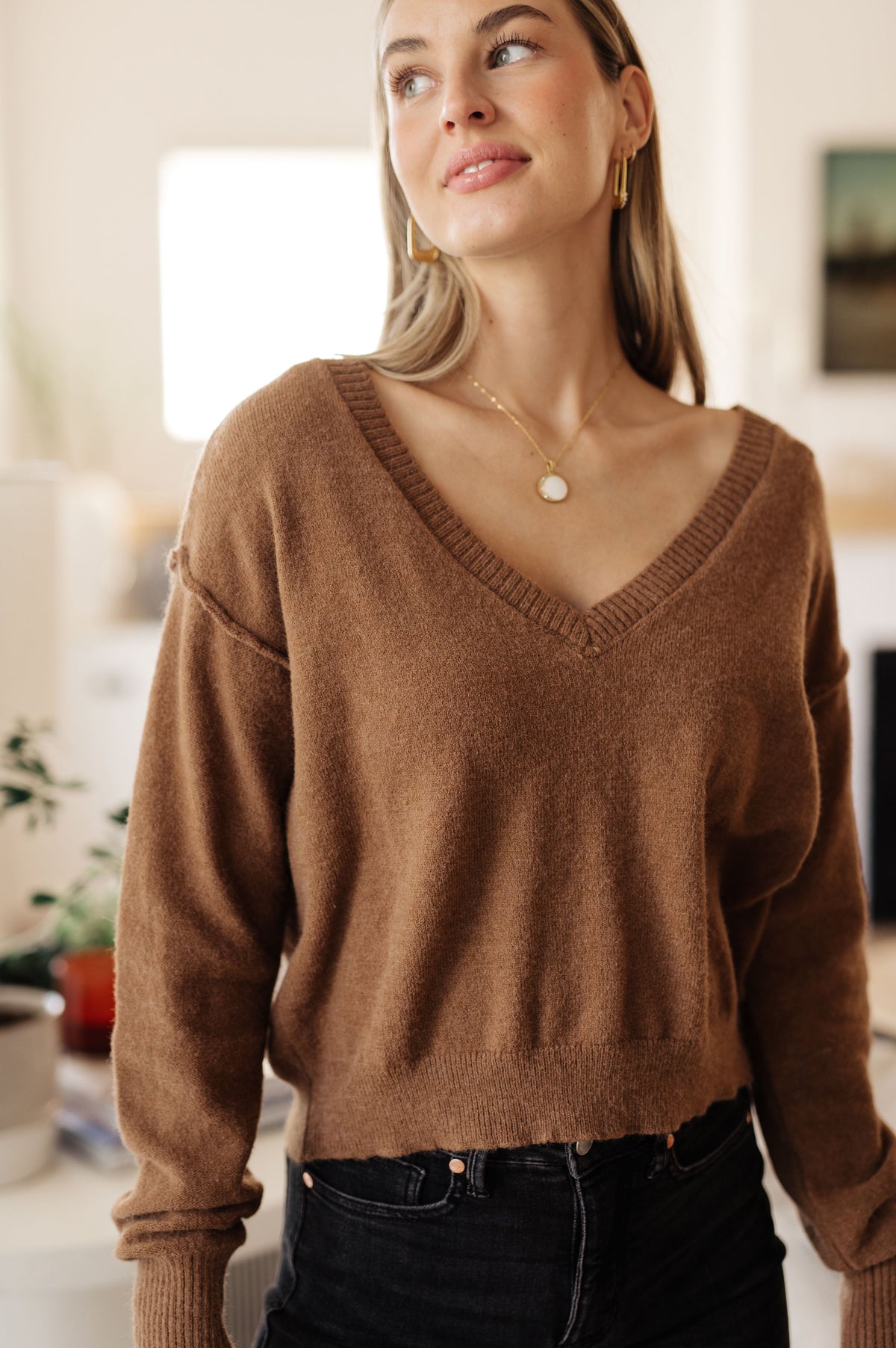 Woman wearing a mocha cropped V neck sweater with dropped shoulders and relaxed sleeves styled with black jeans indoors