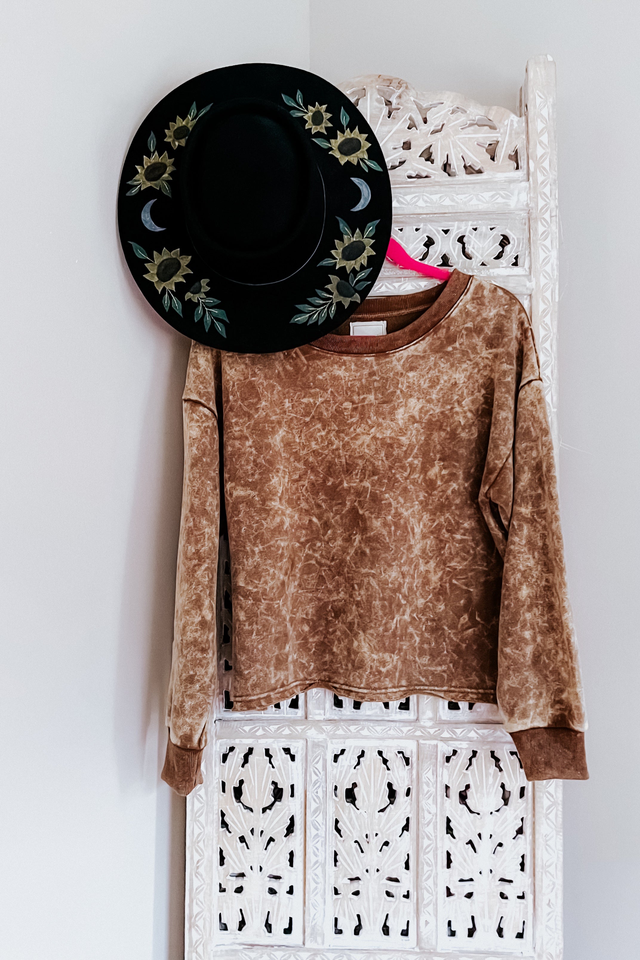 Mocha acid wash cropped pullover hanging on carved white wooden panel with black hat featuring sunflowers and crescent moons