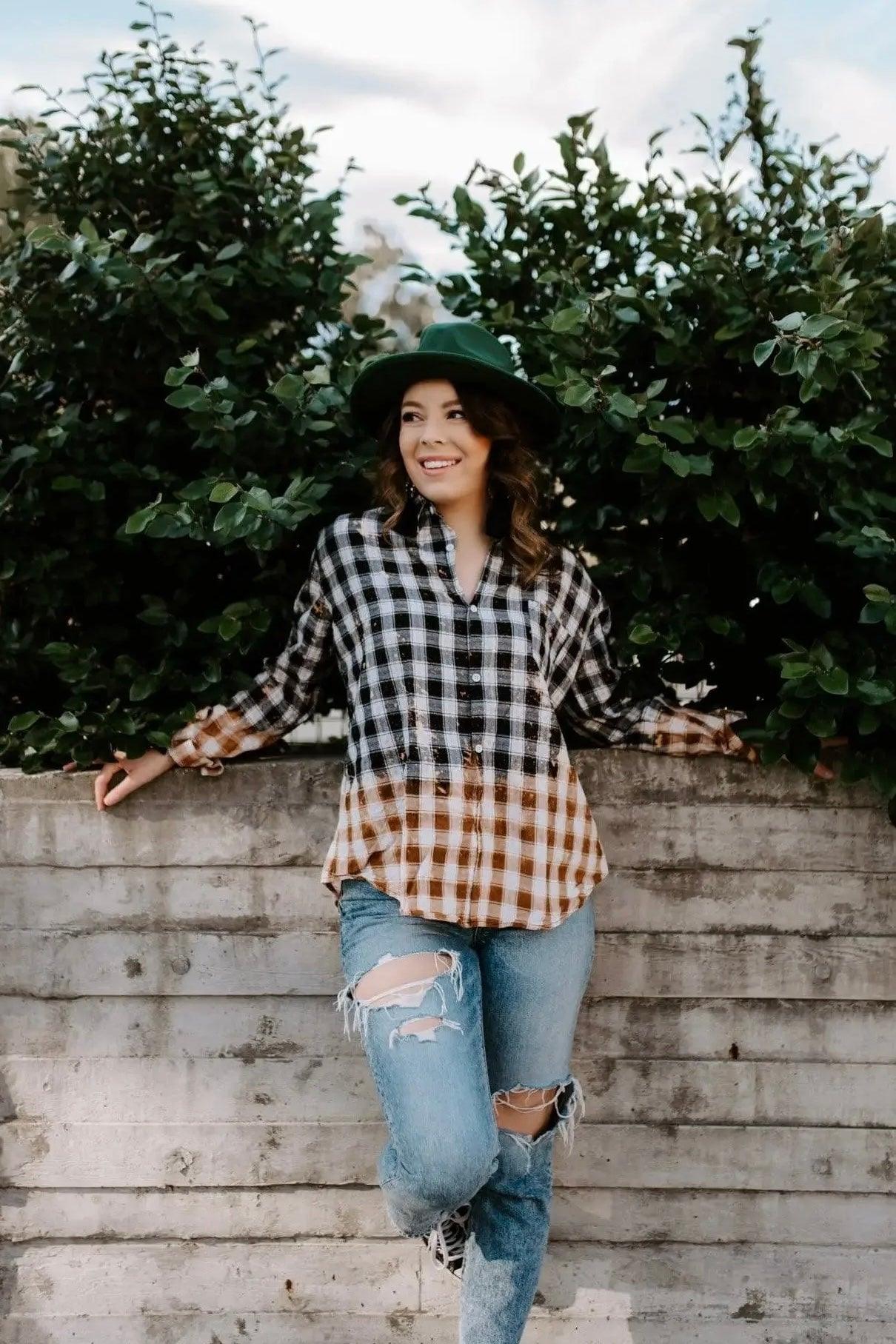 Woman wearing bleach-dyed black and brown plaid flannel with ripped jeans leaning against concrete wall outdoors