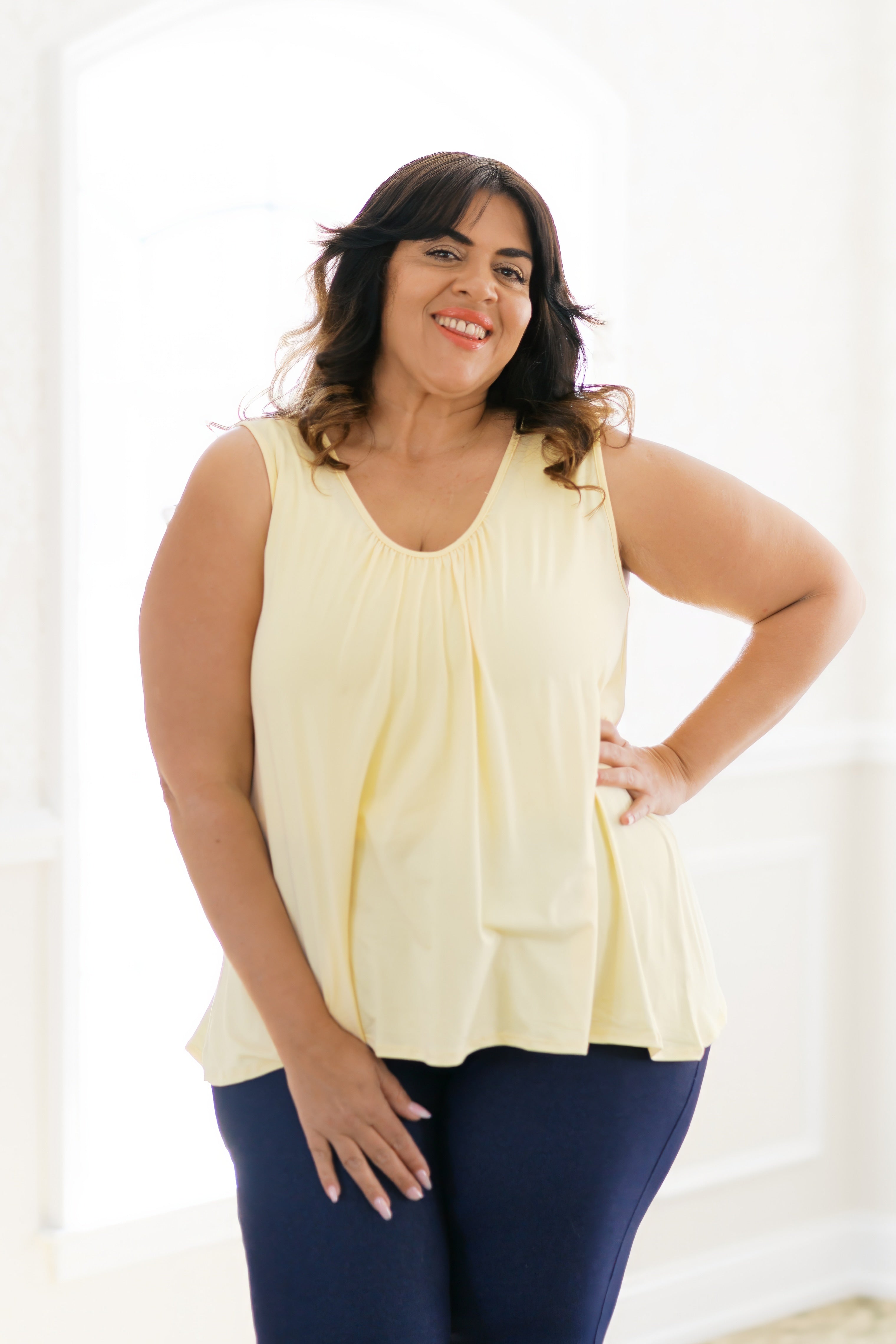 Smiling woman wearing pale yellow Banana Bliss tank top with gathered neckline and navy pants indoors.