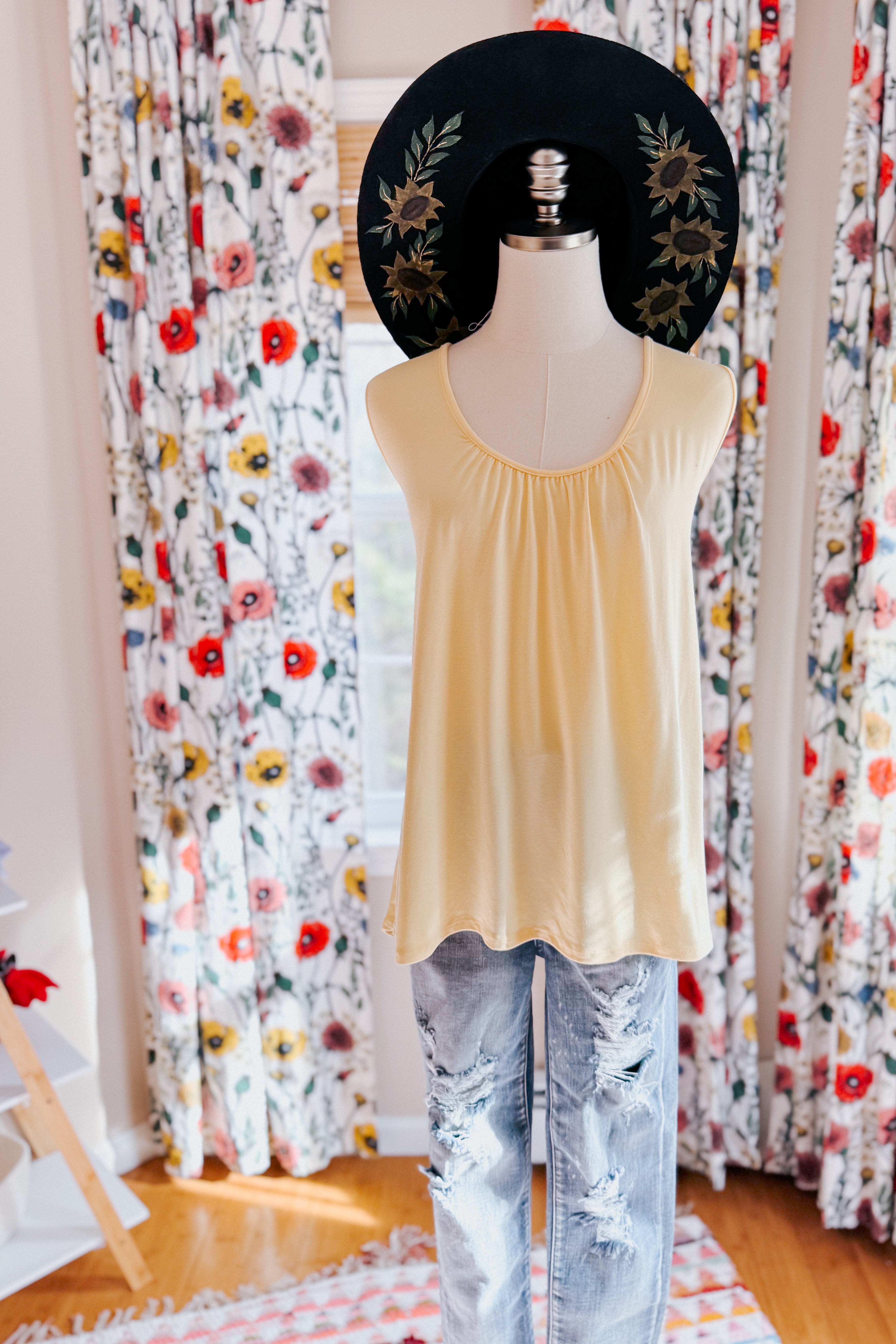 Mannequin dressed in soft yellow Banana Bliss tank top paired with distressed jeans and black floral embroidered wide-brim hat indoors