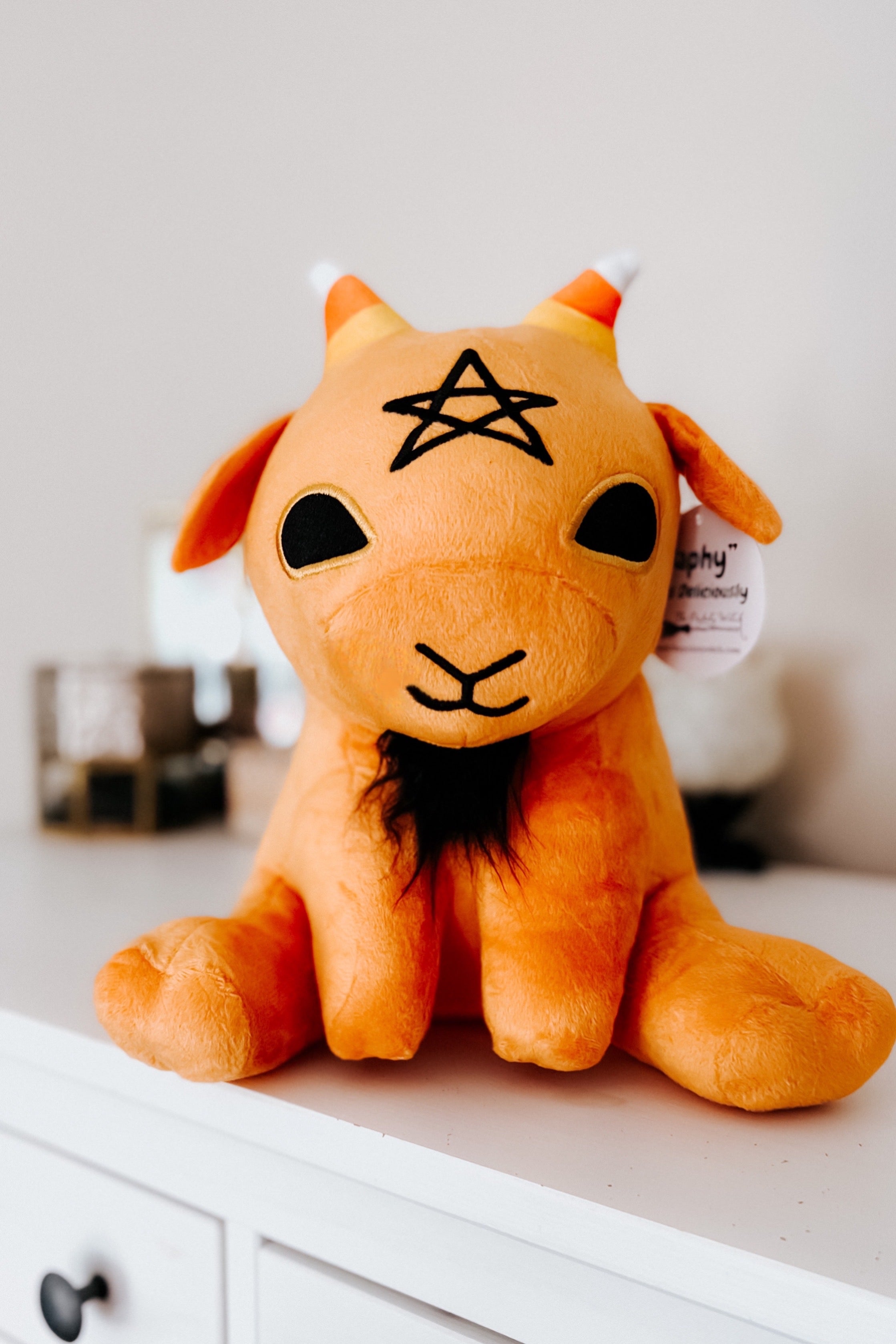 Front view of pumpkin spice Baphy Witchy Baby Goat plush with embroidered star and black beard on white dresser