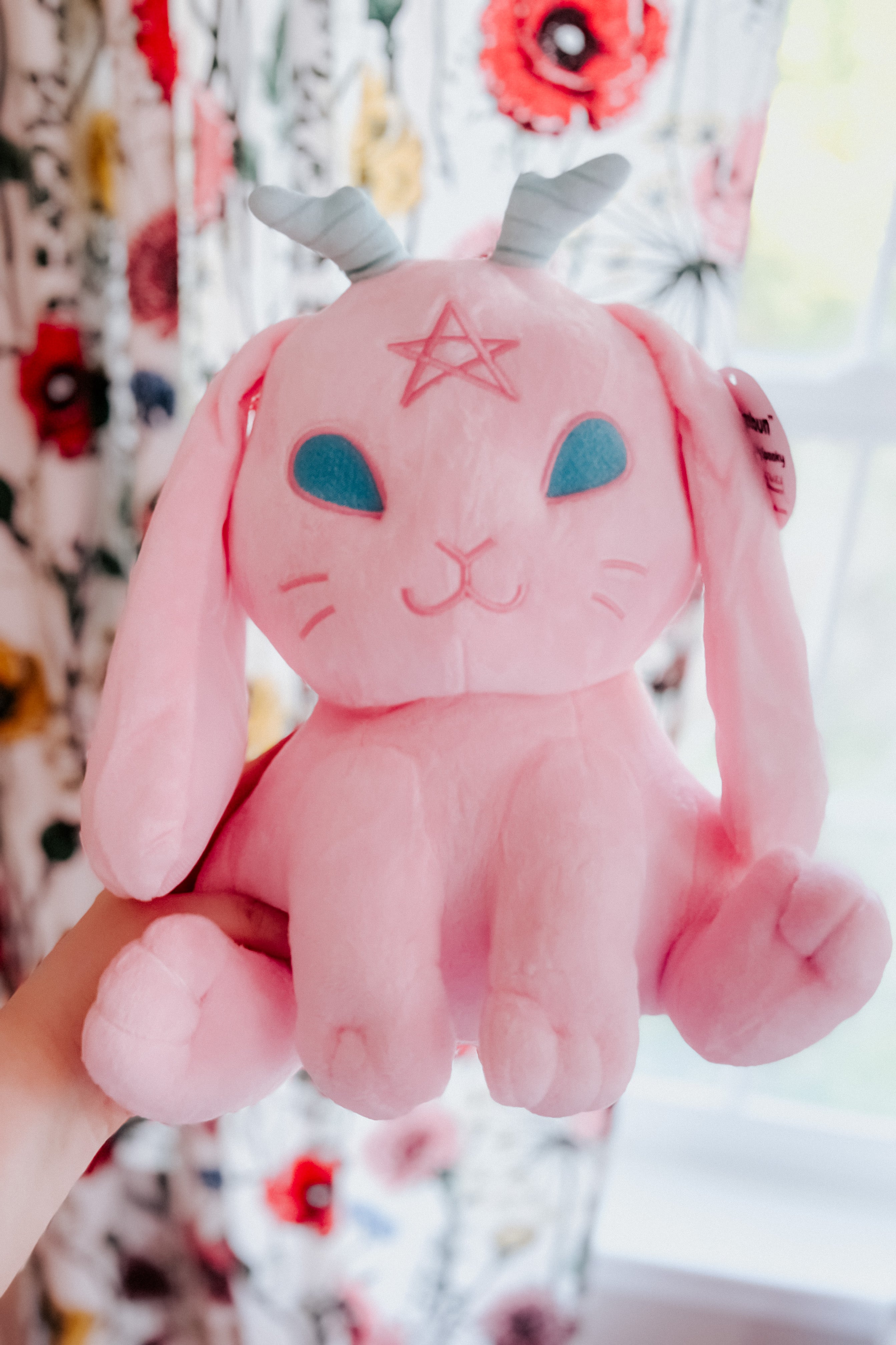 Front view of cotton candy pink Beelzebun Witchy Bunny plush with blue eyes and spiral horns held indoors near floral curtains