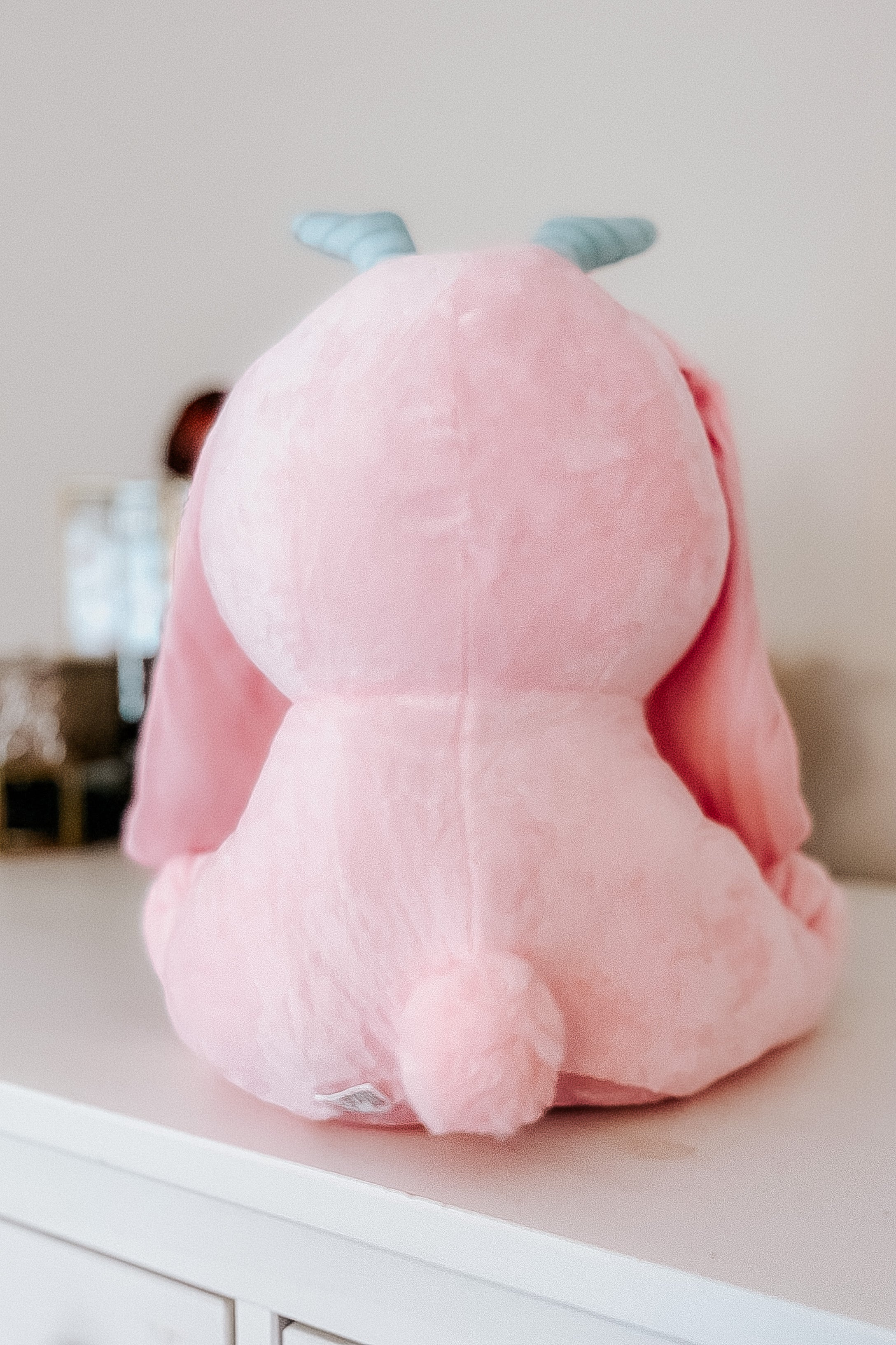 Back view of soft cotton candy pink Beelzebun Witchy Bunny plushie showing tail and blue horns on white surface