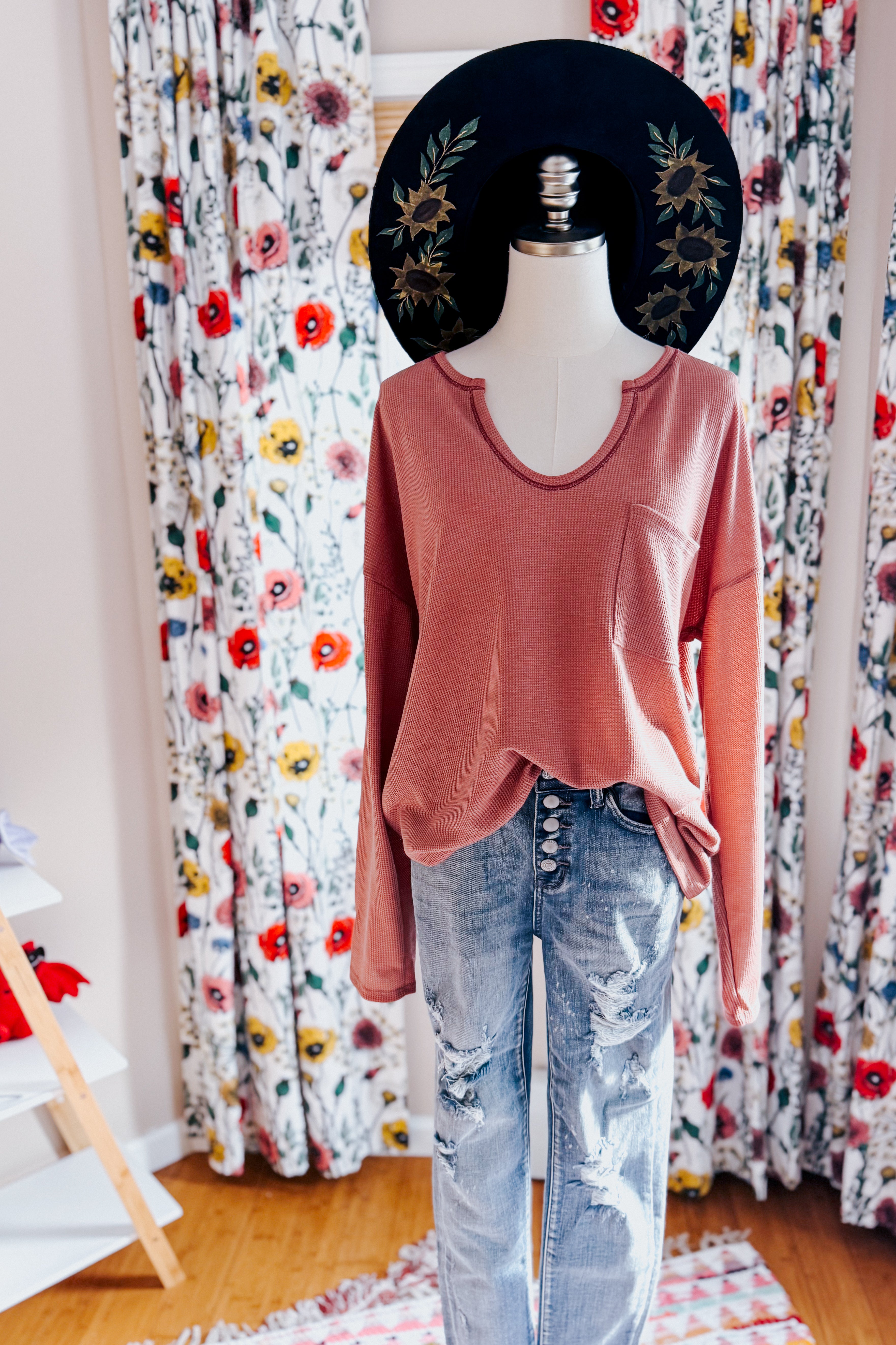 Mannequin styled with rust-colored waffle knit top with pocket and distressed button-fly jeans in floral curtain backdrop