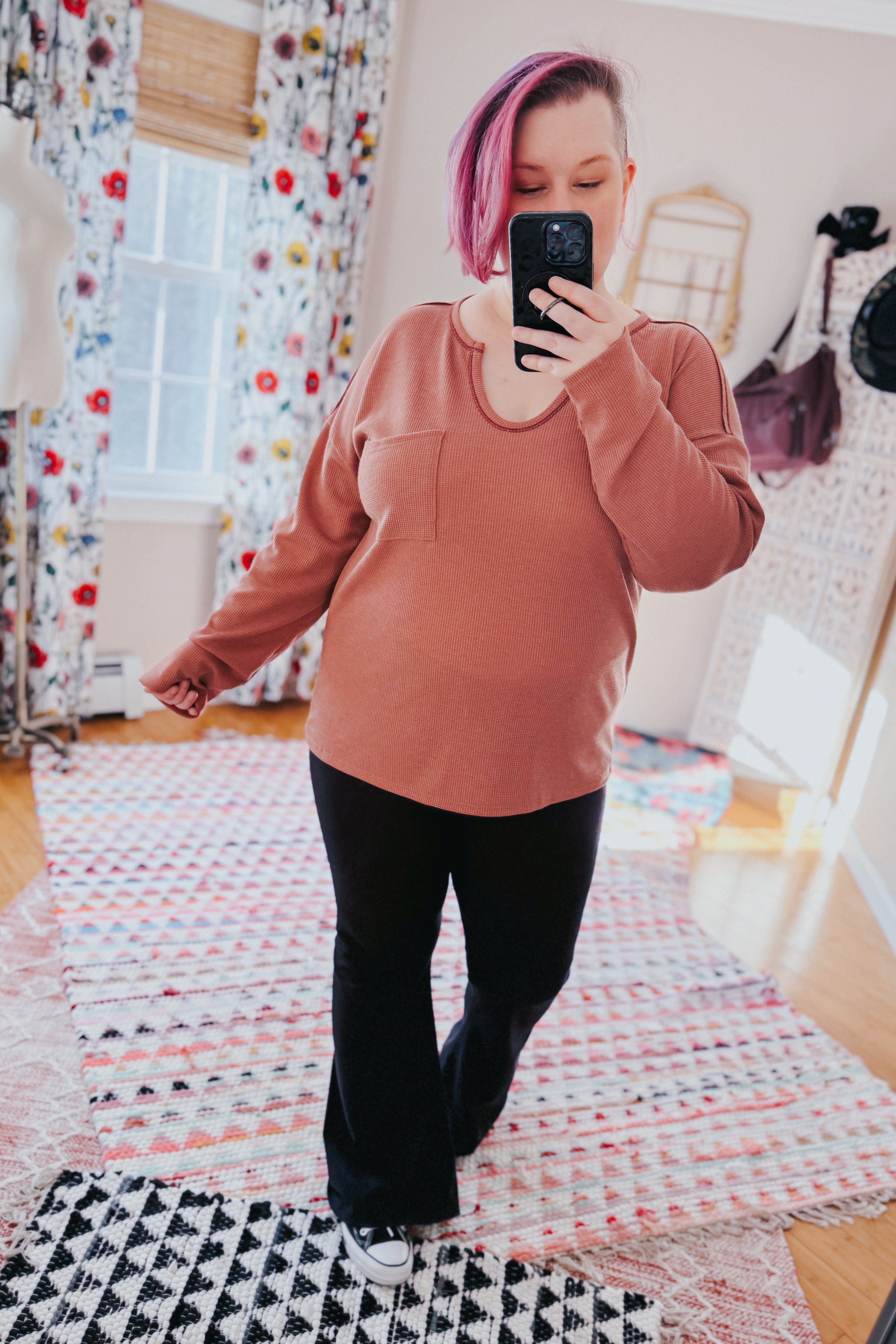 Individual wearing rust-colored Behind The Scenes waffle knit top with pocket taking mirror selfie in colorful room with vibrant curtains