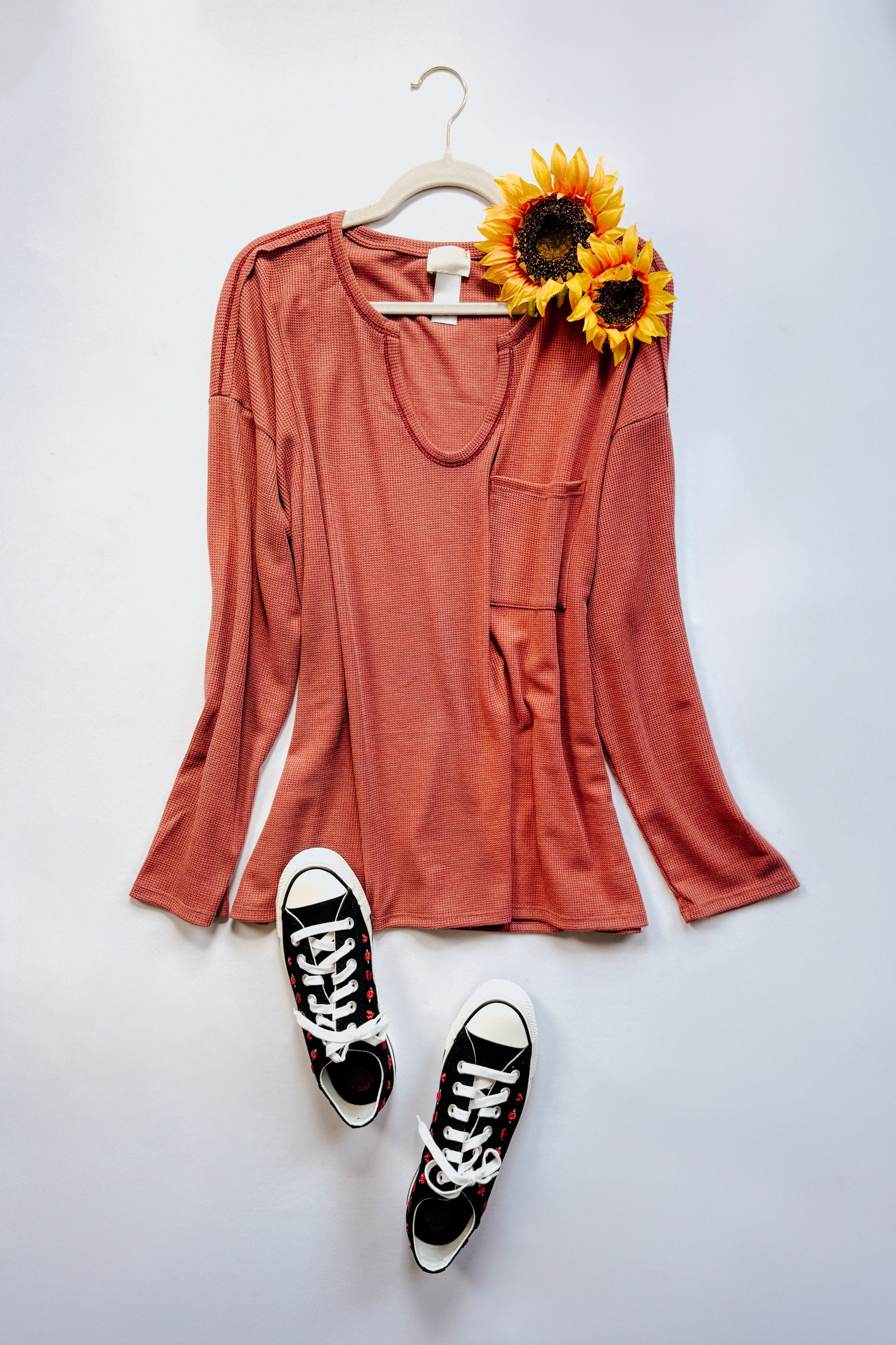 Rust-colored waffle knit top with front pocket displayed with black and white sneakers and two sunflowers on white background