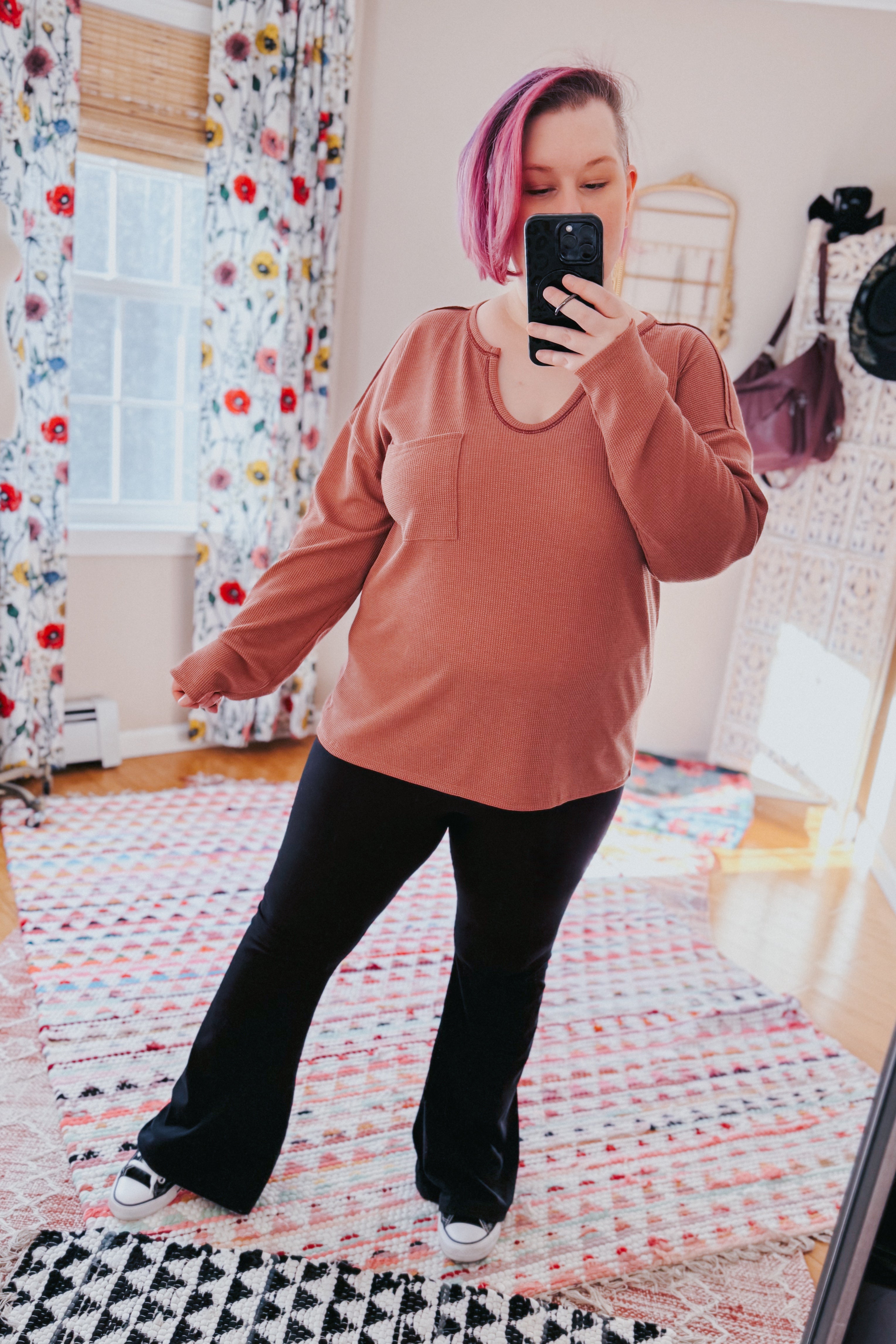 Woman with pink hair taking mirror selfie wearing rust-colored waffle knit top with pocket and black pants in sunlit room