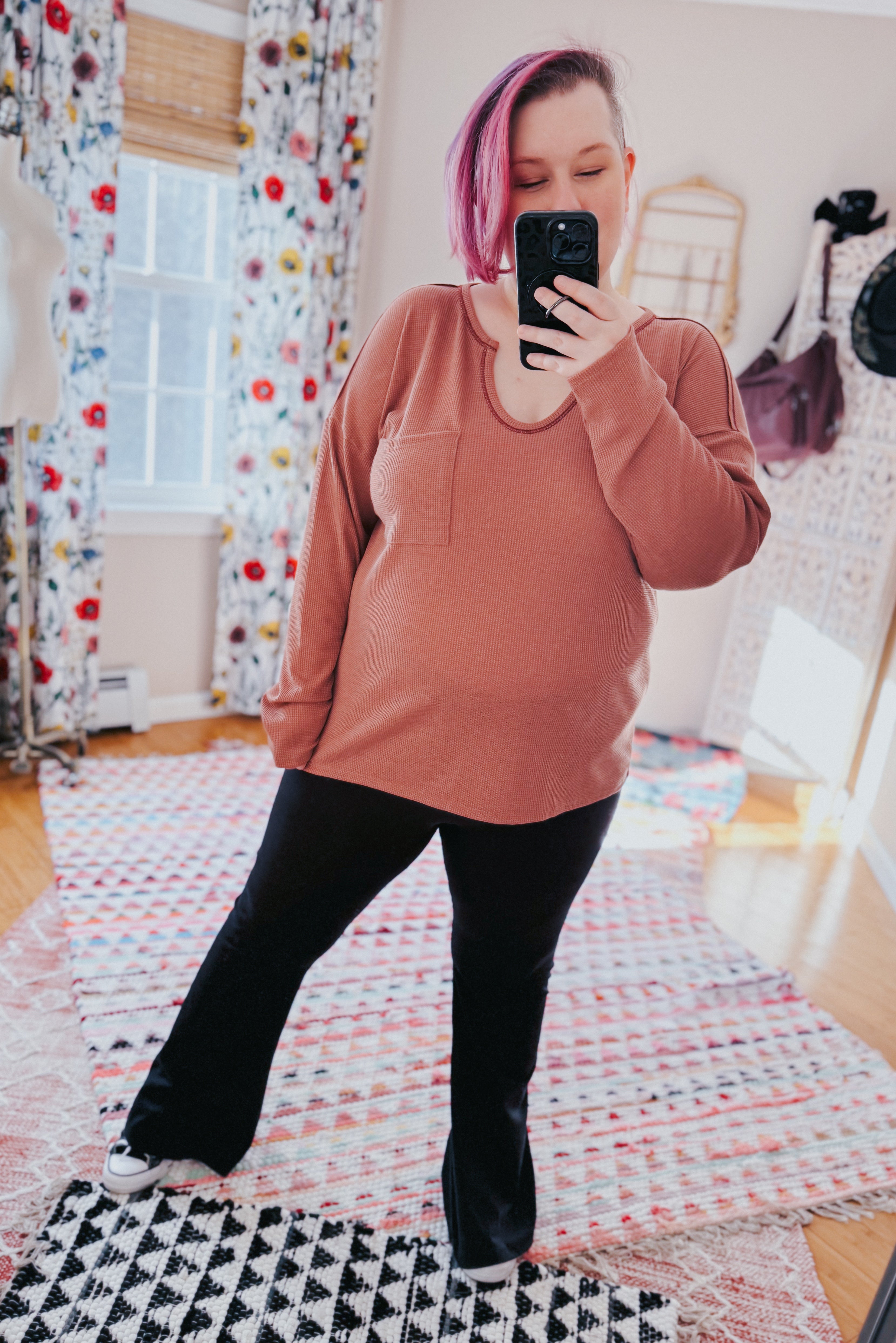 Woman taking mirror selfie wearing rust-colored waffle knit top with pocket and black pants in bright room