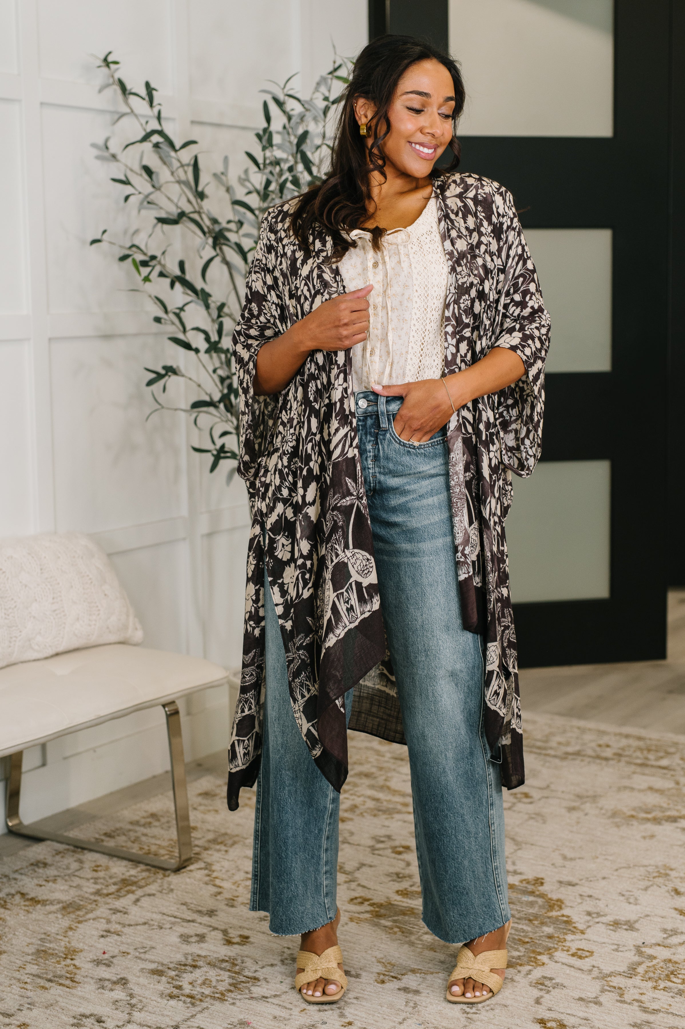Smiling woman wearing black floral print duster over white top and wide-leg jeans in cozy indoor setting