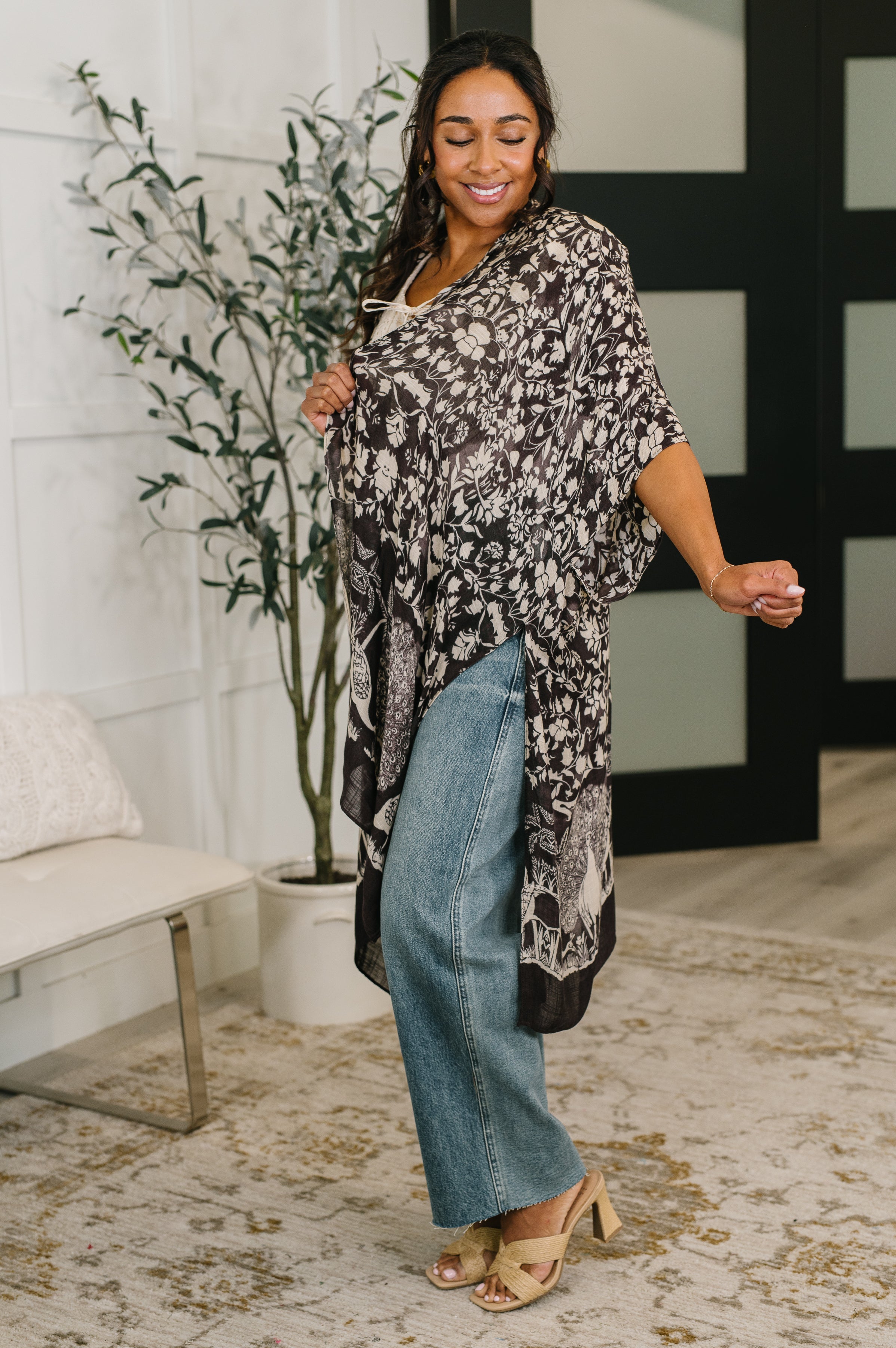 Woman wearing black floral open-front duster over jeans and tan heeled sandals in stylish indoor space
