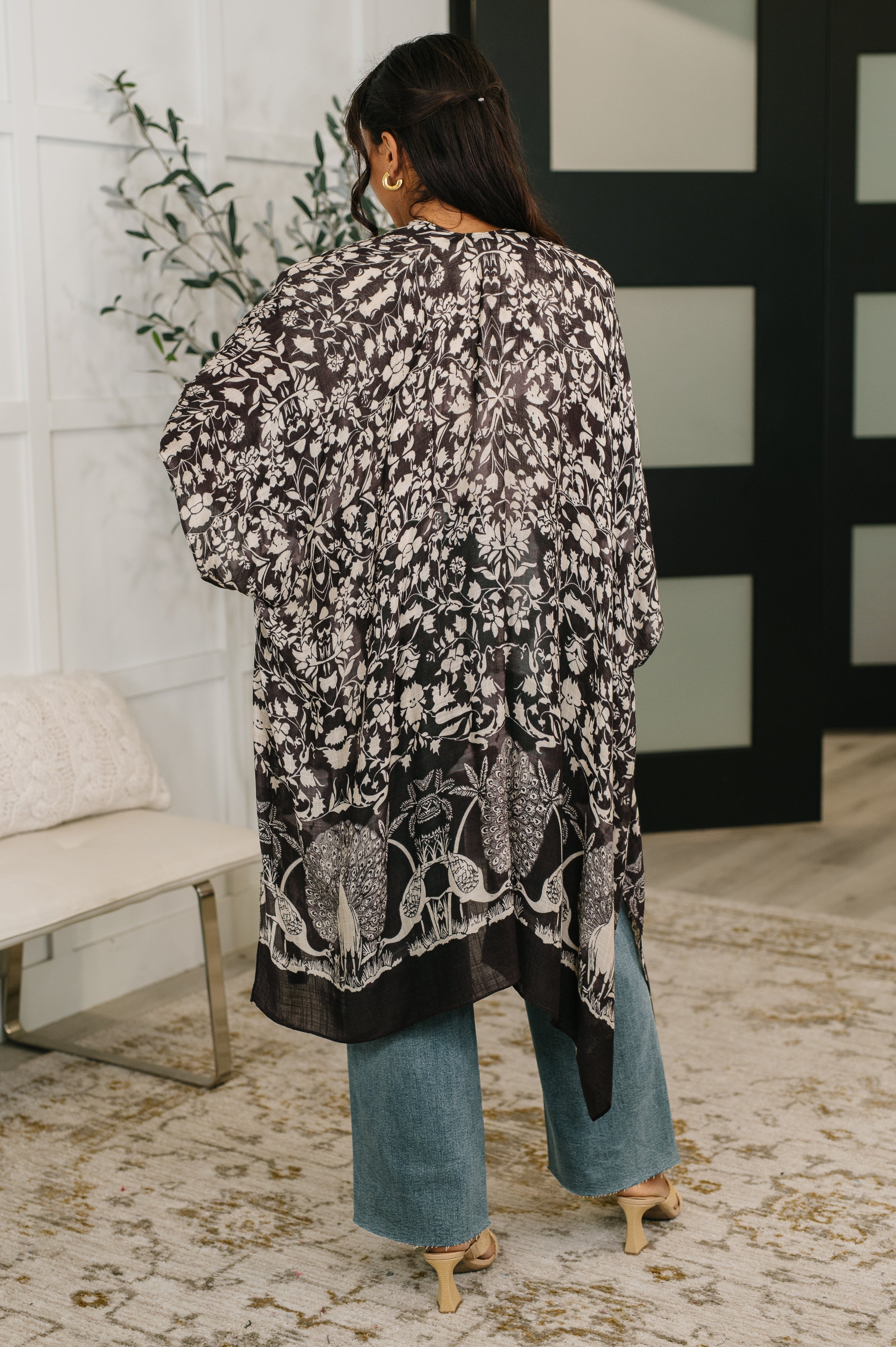 Back view of woman wearing black floral print open front duster over wide-leg jeans and heels indoors