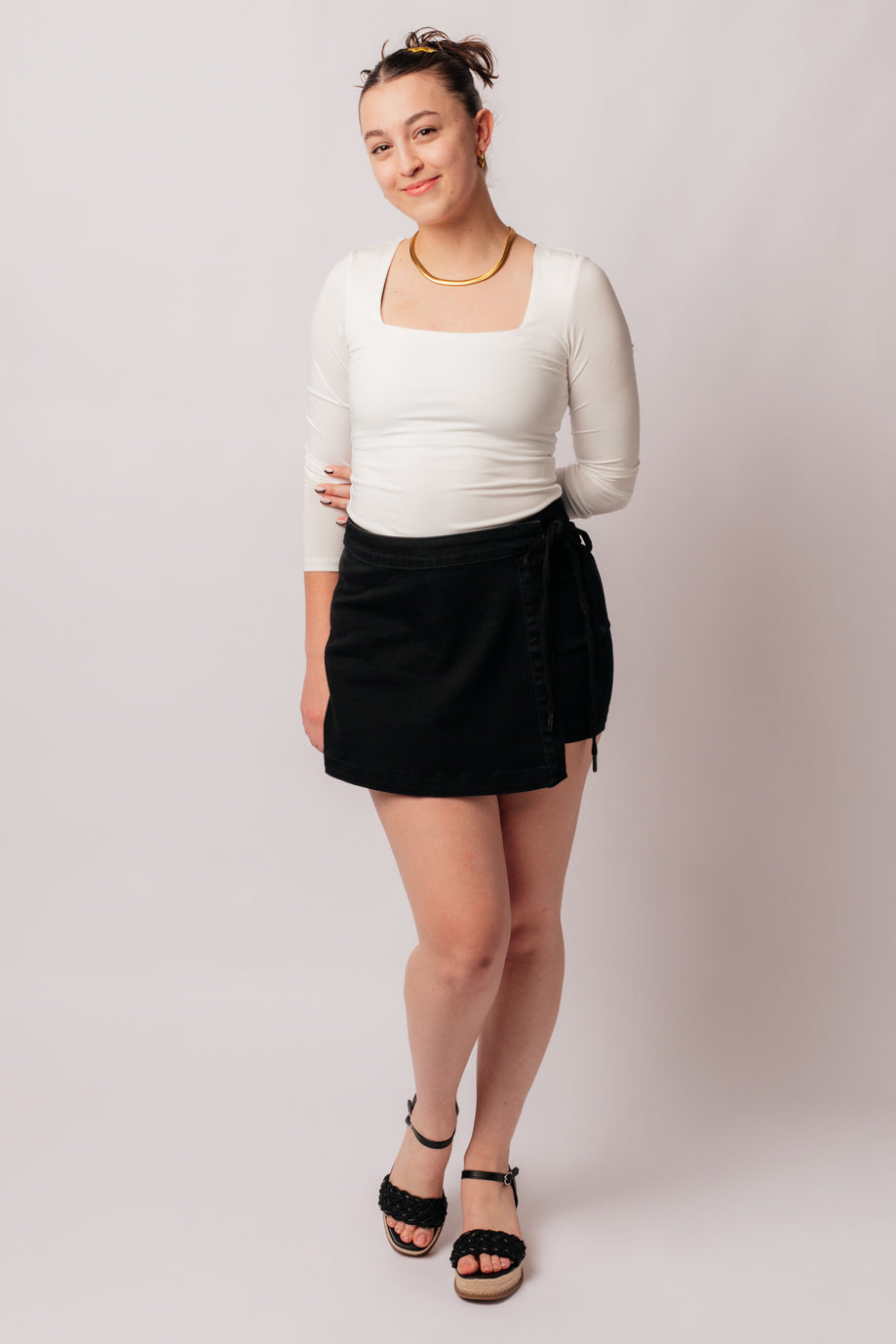 Woman wearing Bentli black high rise denim skort with side tie detail paired with white square neck top and black sandals