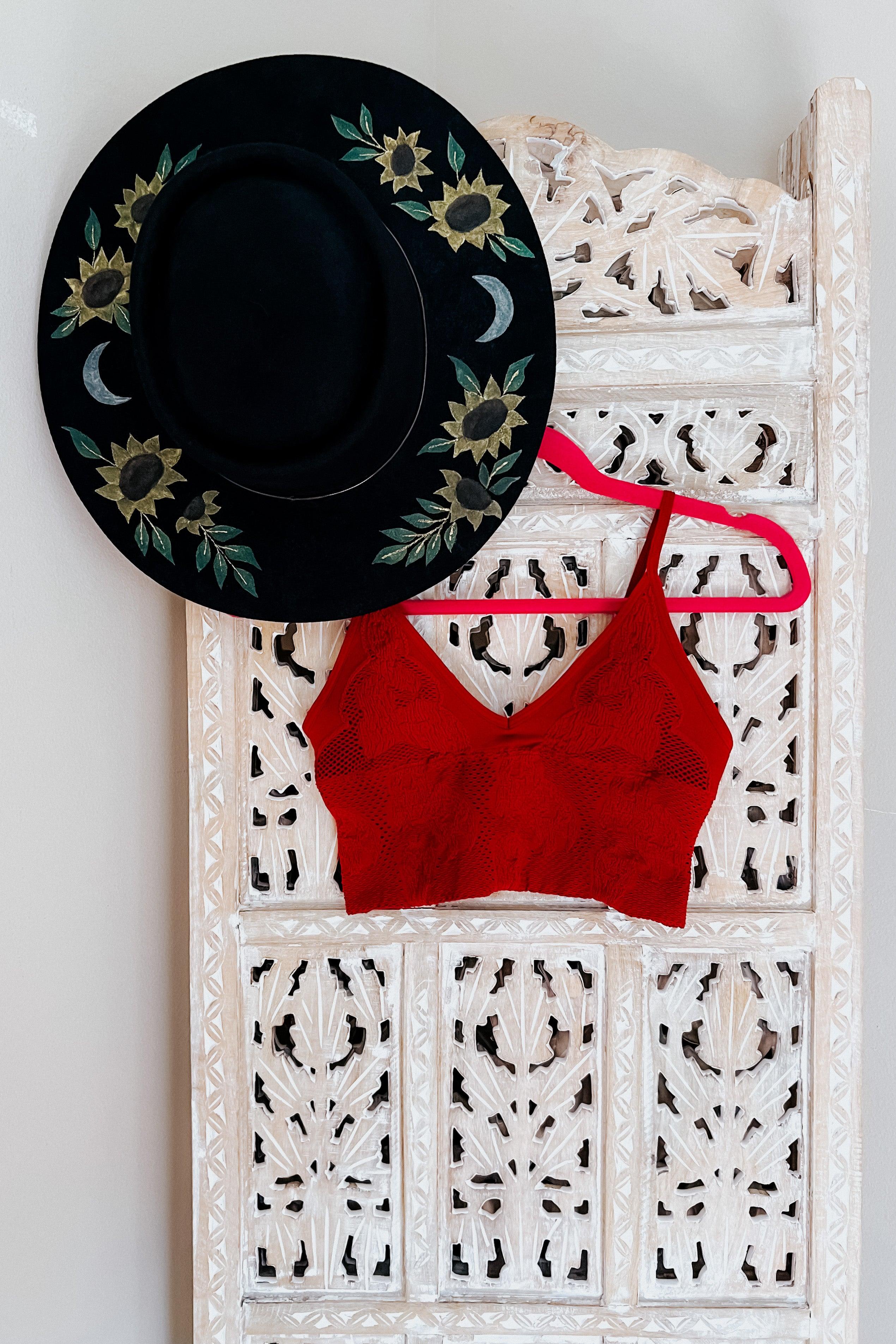 Red seamless padded textured brami with thin straps hanging on pink hanger next to black hat with floral and moon embroidery
