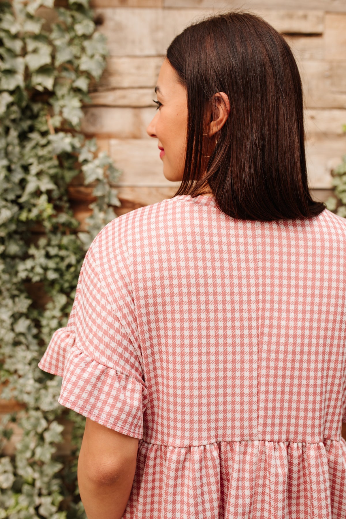 Woman seen from behind wearing pink and white gingham dress with ruffle sleeves standing near ivy-covered wall