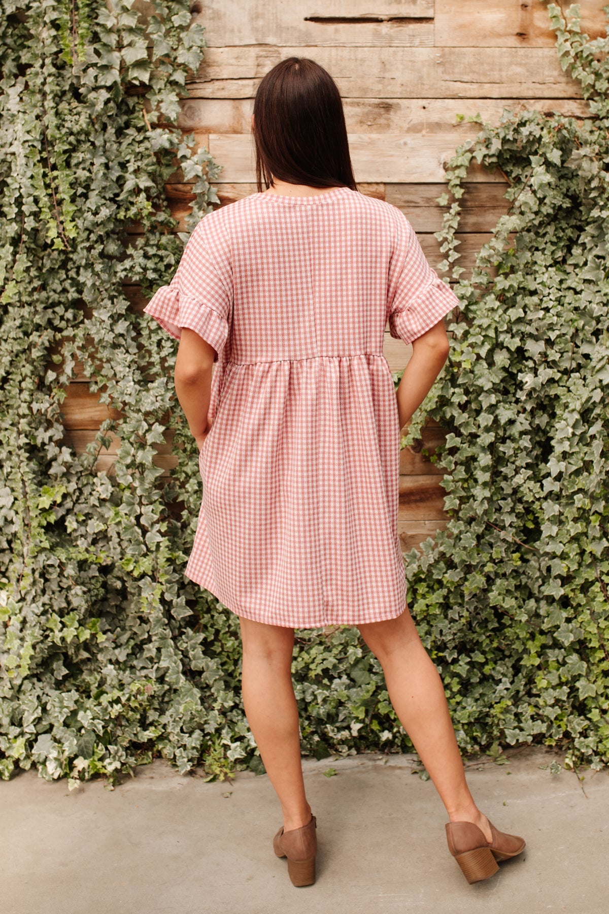 Woman wearing pink gingham short sleeve dress with ruffle detail and brown block heel shoes standing outdoors with ivy backdrop