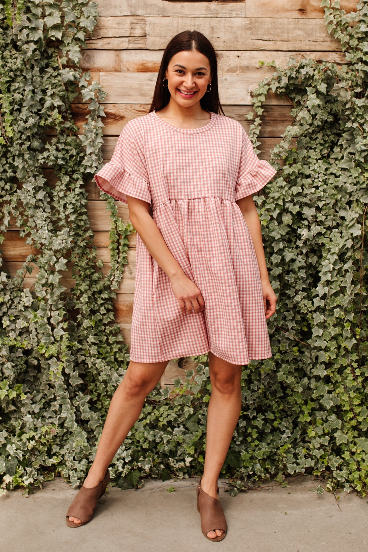 Woman smiling in pink gingham babydoll dress with ruffled sleeves and brown peep-toe heels by ivy-covered wood wall